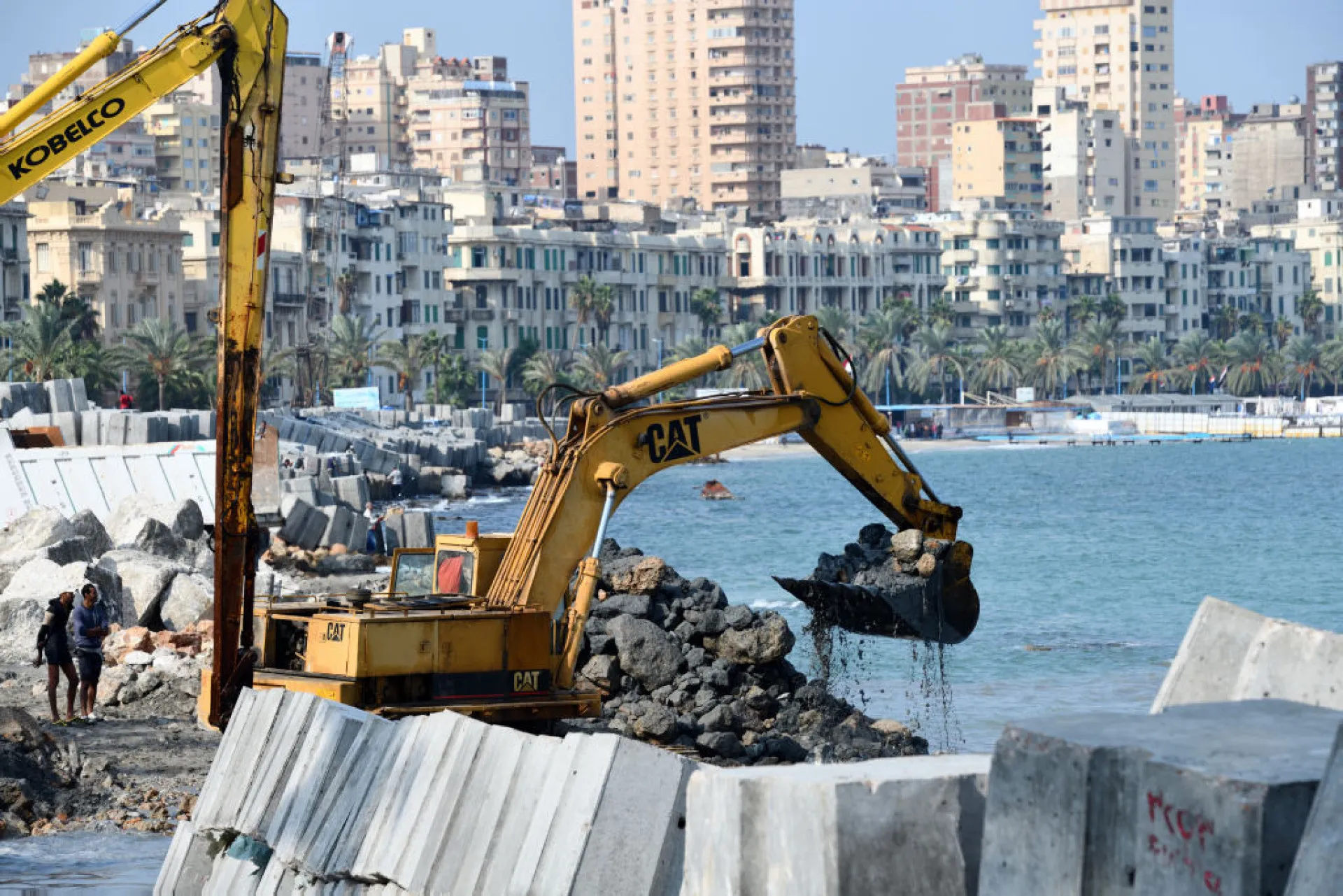 Pessoas caminhando na orla de Alexandria, com o mar calmo ao fundo, em um momento de aparente tranquilidade.