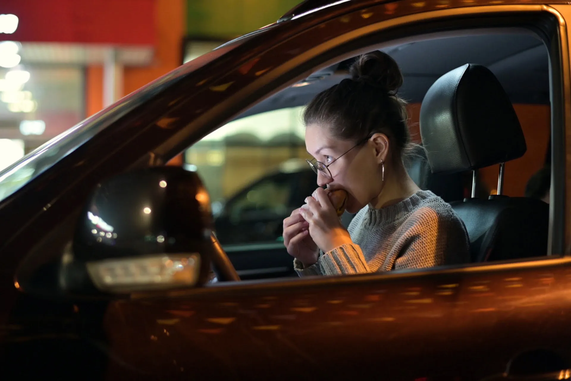 Uma pessoa comendo um hambúrguer e batatas fritas dentro do carro, uma cena comum para quem busca uma refeição rápida.