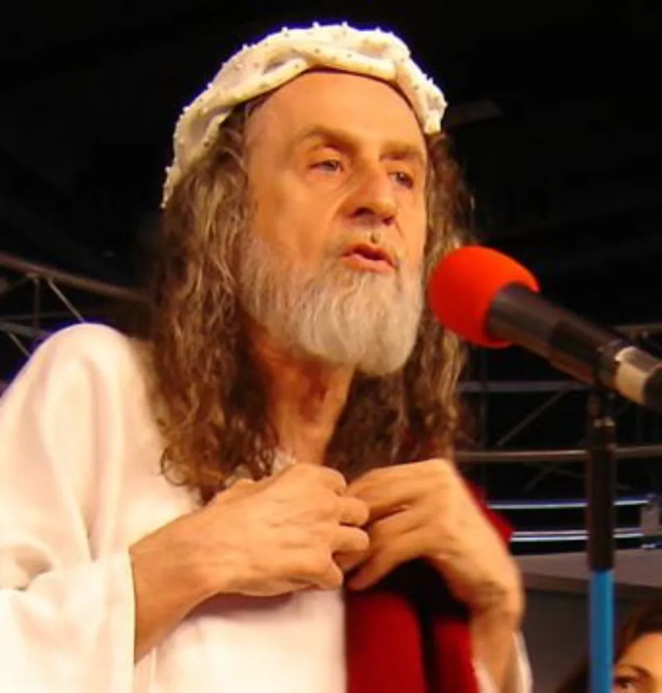 Inri Cristo, sentado em um trono, vestido com uma túnica branca e coroa, cercado por suas seguidoras.