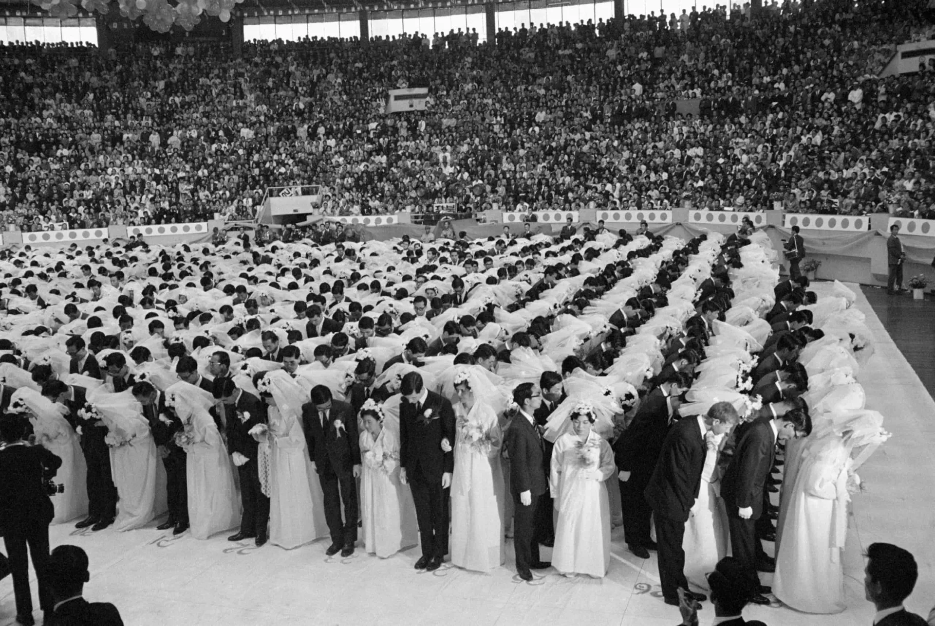 Milhares de casais vestidos com trajes de casamento idênticos em um estádio, durante uma cerimônia da Igreja da Unificação.