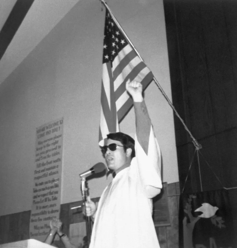 O Reverendo Jim Jones falando em um microfone, com seus característicos óculos escuros e expressão intensa.