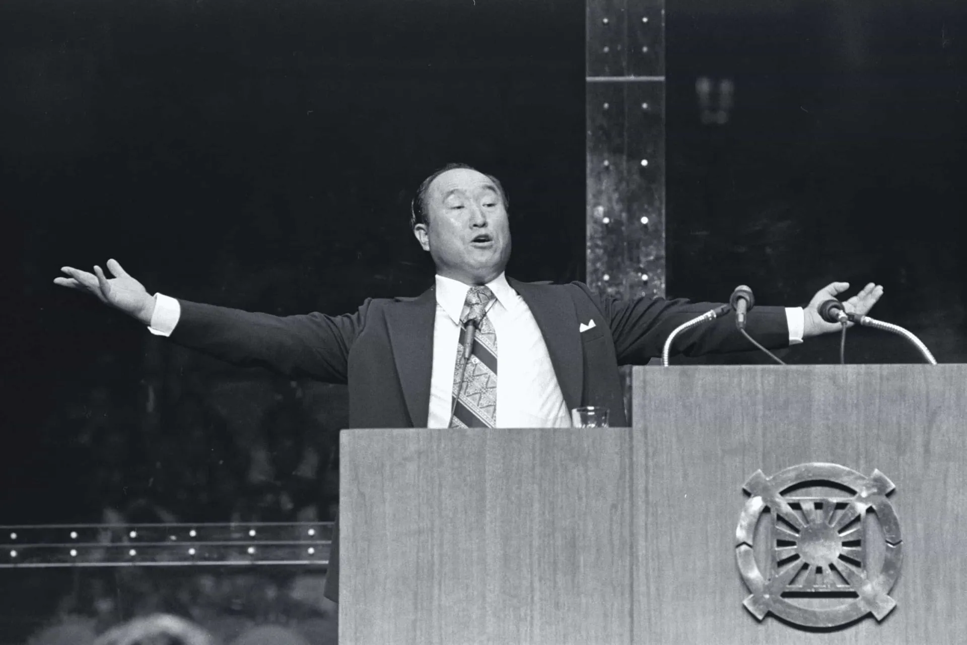 Sun Myung Moon discursando em um grande evento, com uma expressão séria e determinada.
