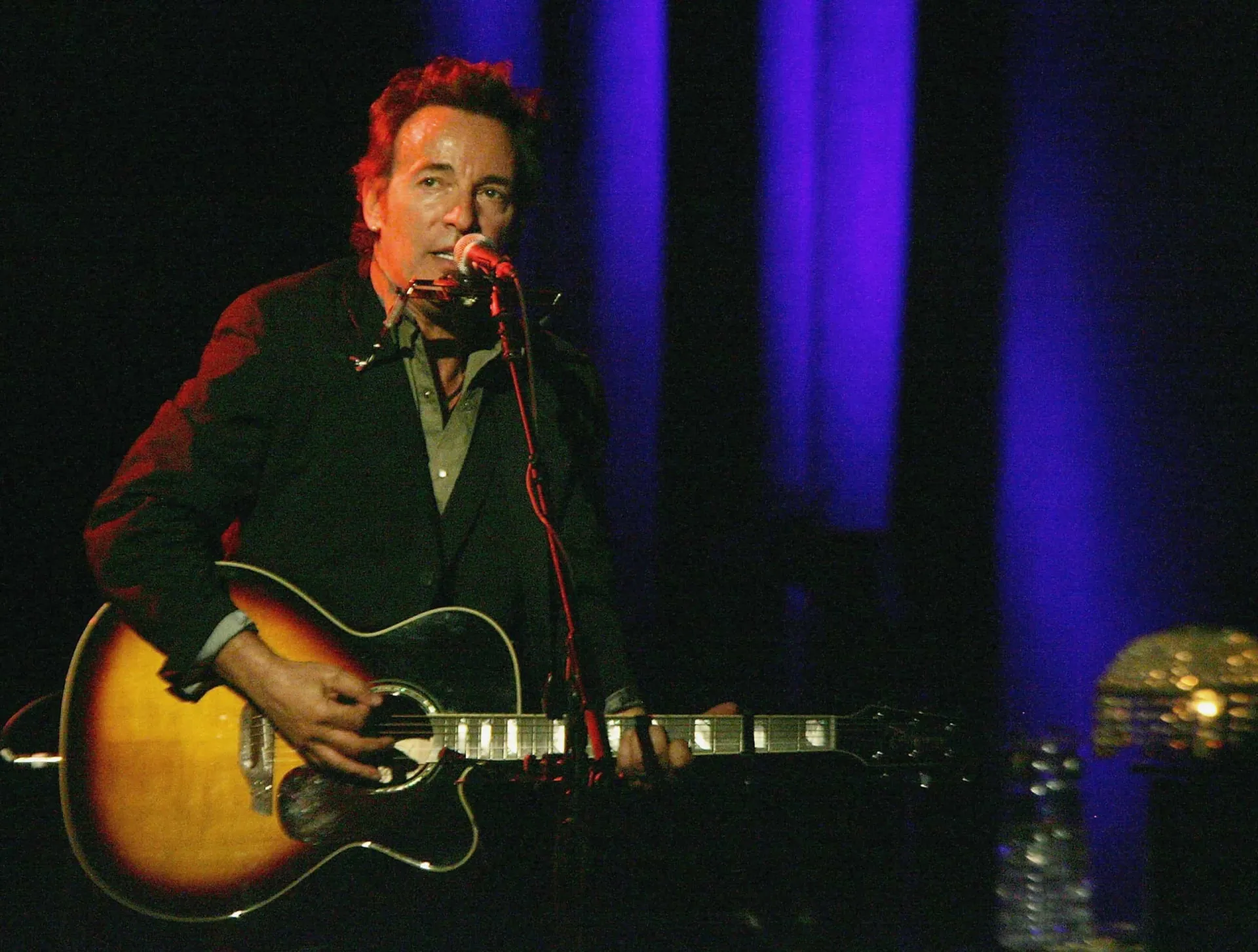 Bruce Springsteen tocando violão e gaita, com uma expressão concentrada.
