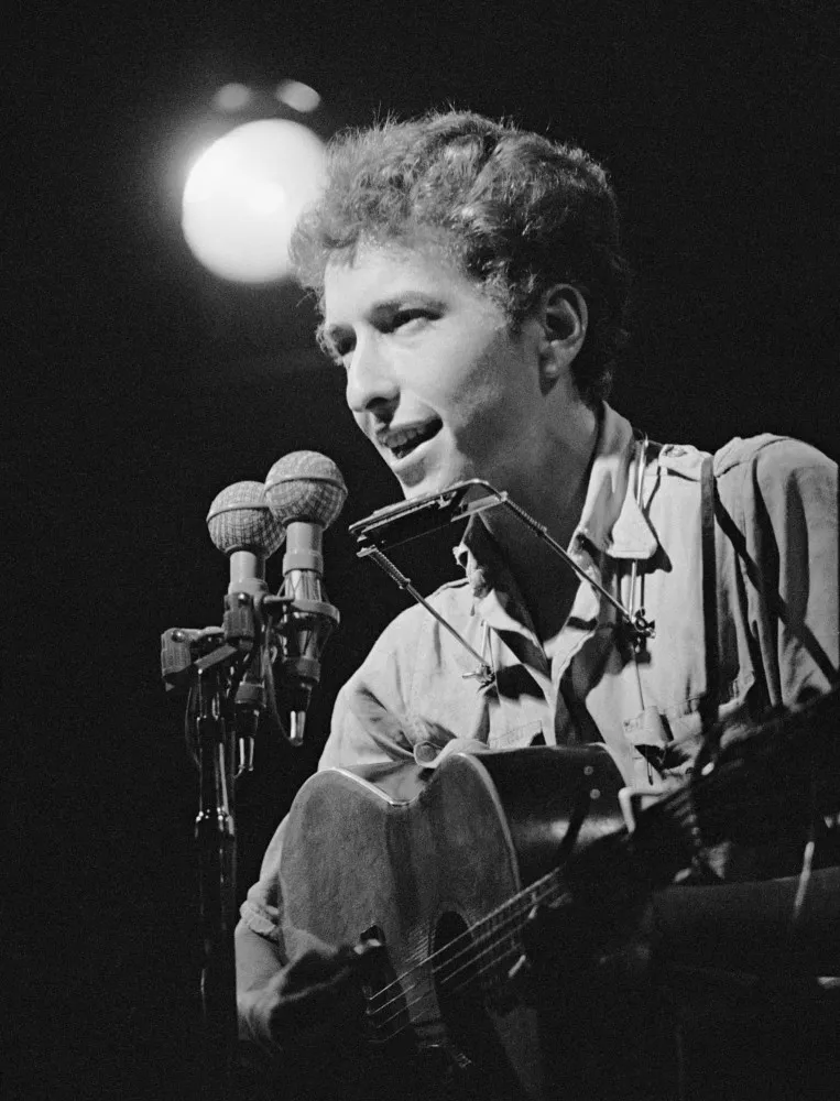 Bob Dylan, jovem, tocando violão e gaita em uma foto clássica em preto e branco.
