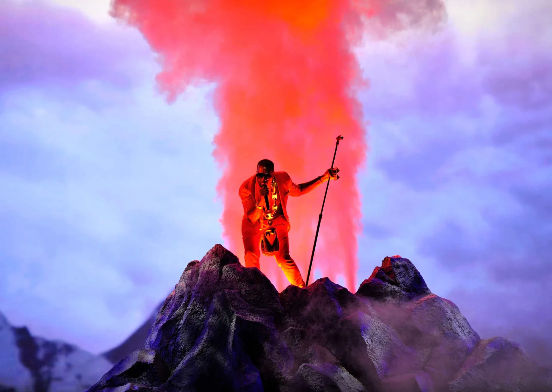 Kanye West no palco, usando óculos escuros e uma jaqueta vermelha vibrante.