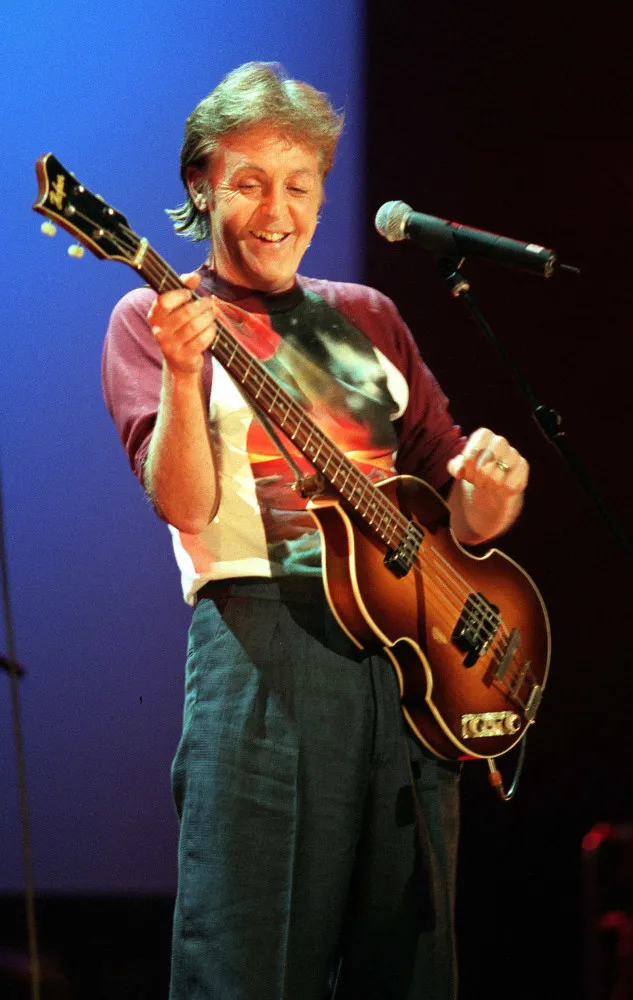 Paul McCartney sorrindo e tocando seu clássico baixo Hofner no palco.