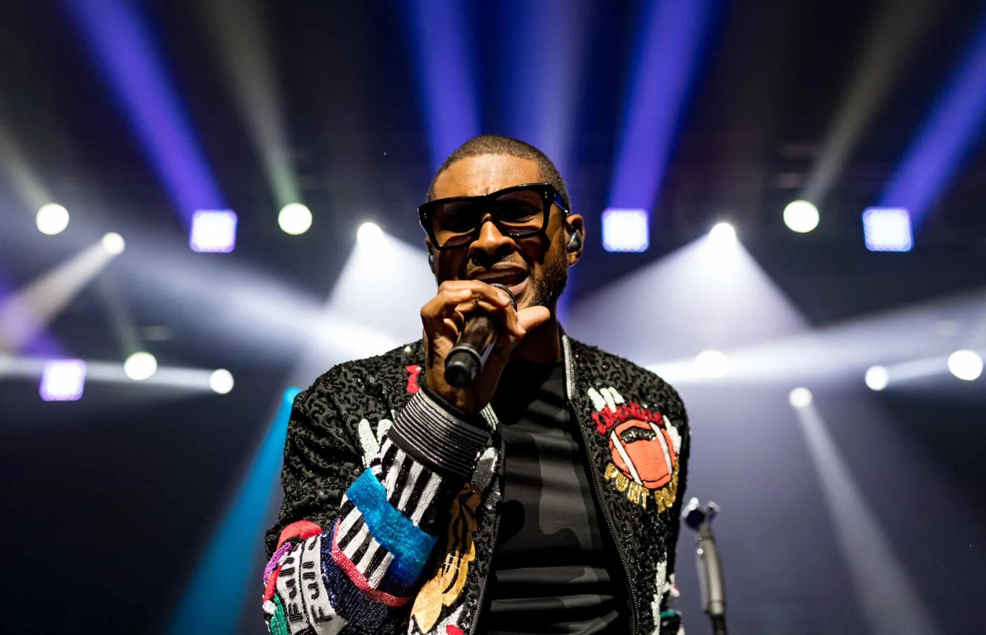 O cantor Usher se apresentando no palco, com um microfone e dançando.