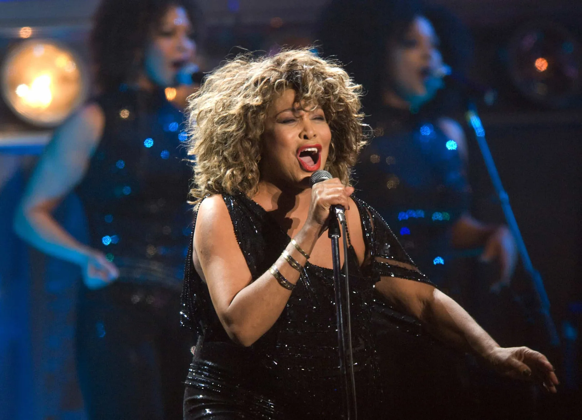 Tina Turner em uma performance energética no palco, com seu cabelo icônico e vestido brilhante.