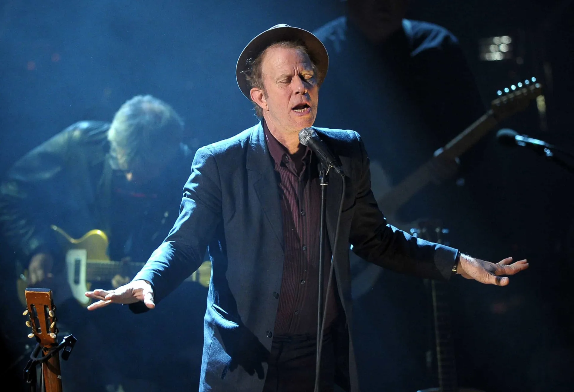 Tom Waits com seu chapéu característico, cantando em um microfone com sua voz rouca.
