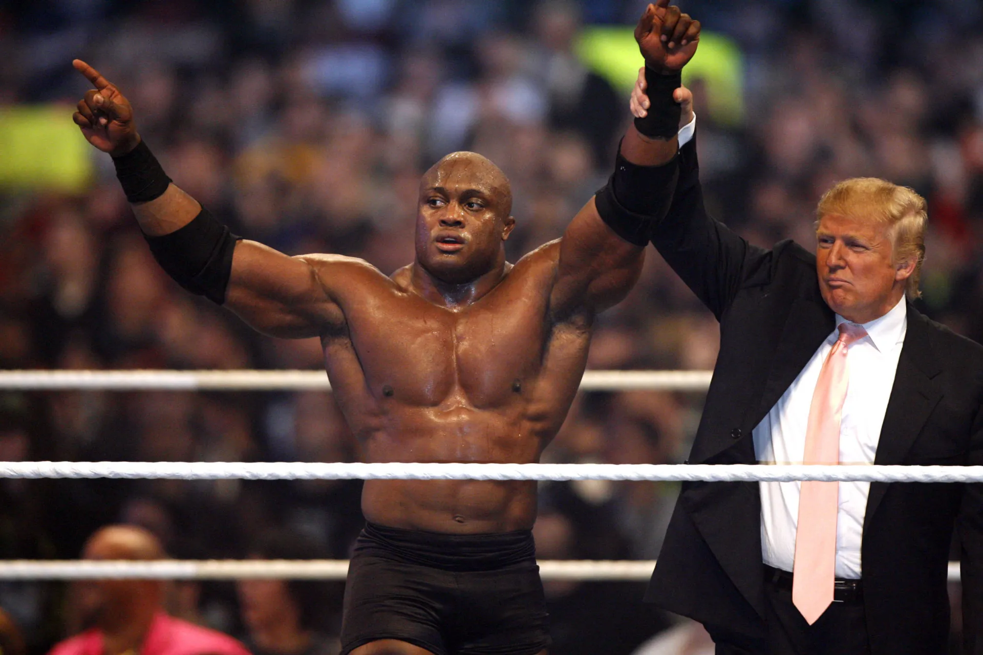 Donald Trump no centro de um ringue da WWE, interagindo com os lutadores.