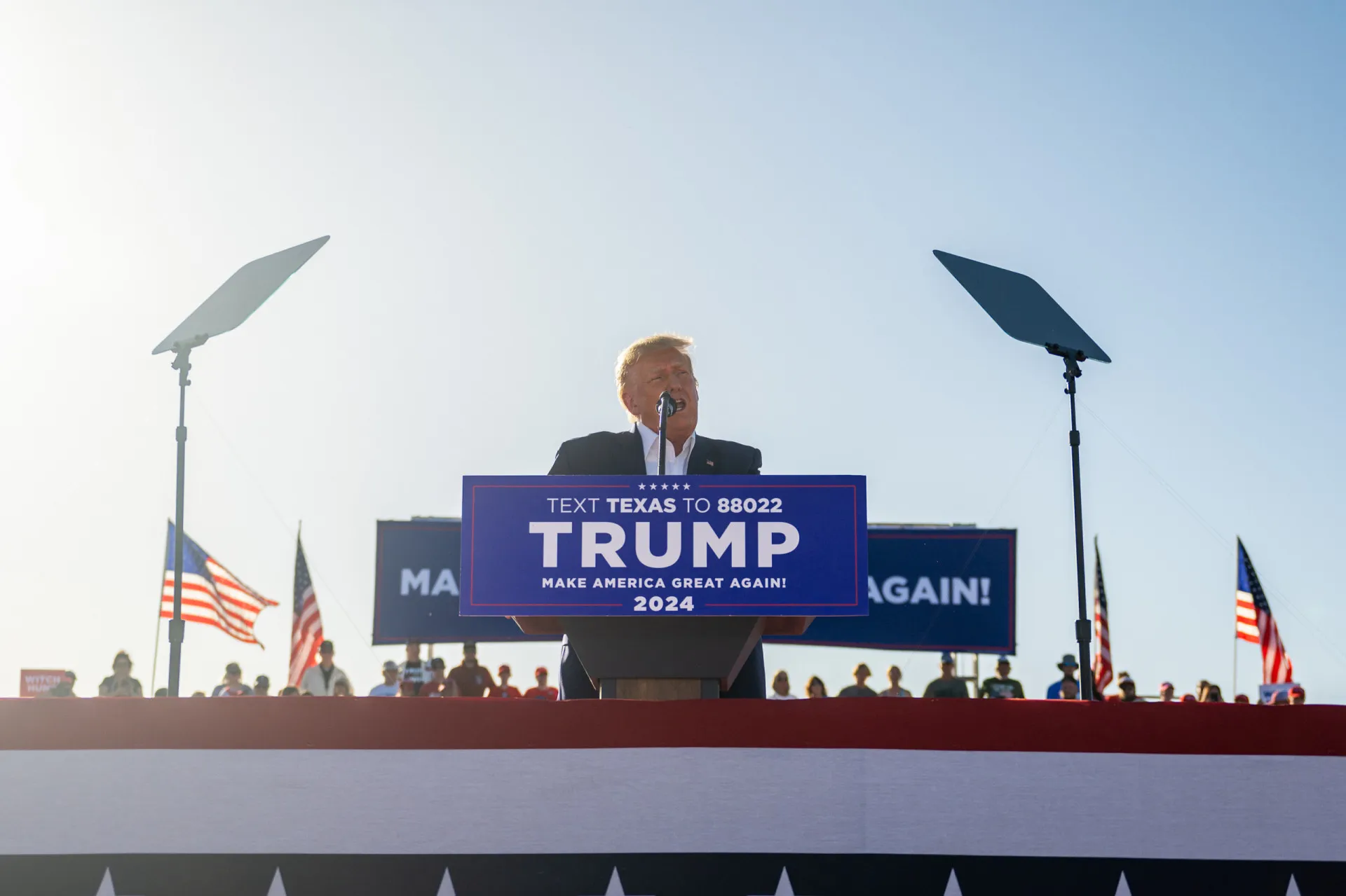 Donald Trump em um comício de campanha, acenando para uma multidão de apoiadores.