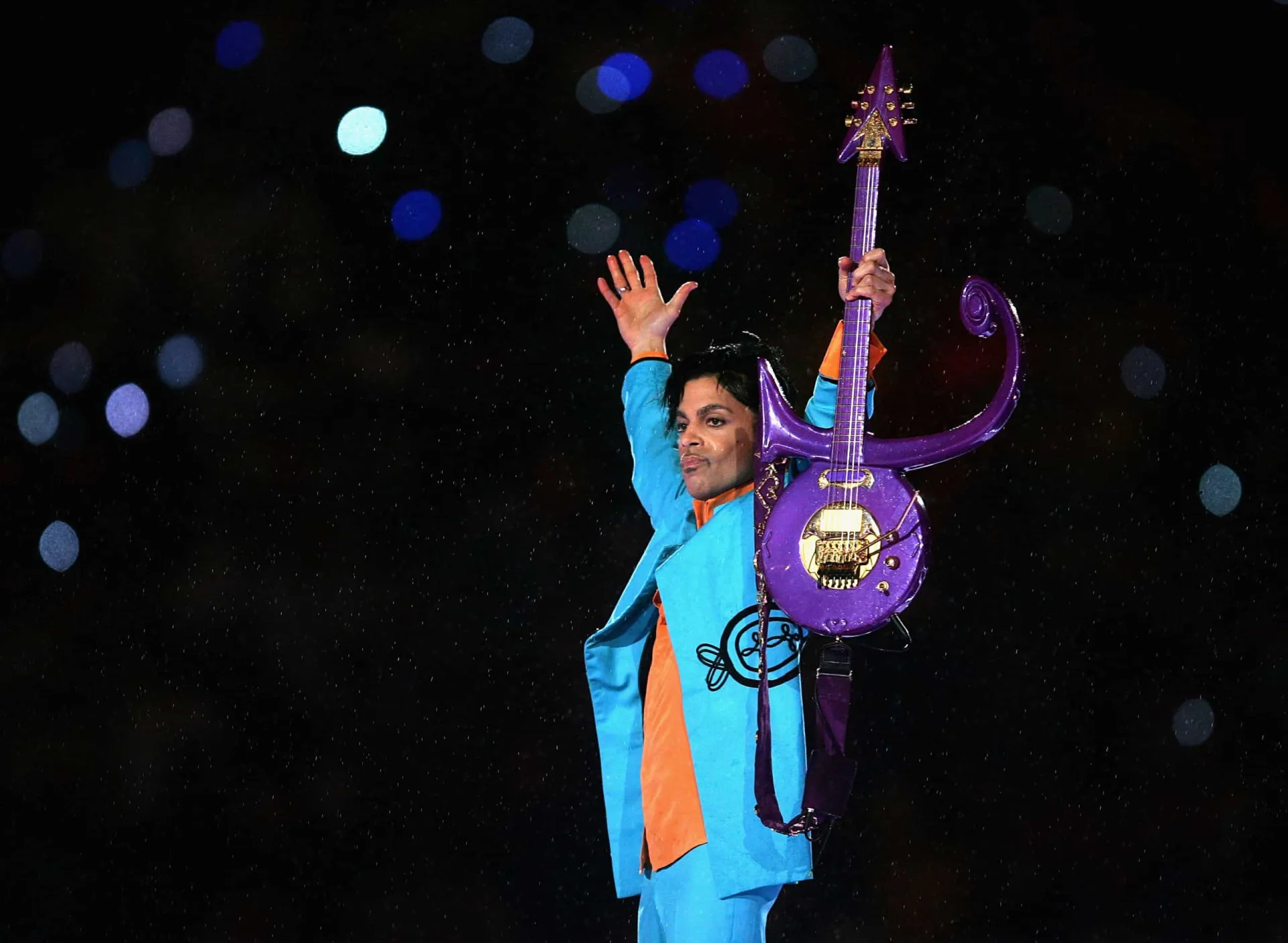 Prince tocando sua guitarra característica no palco, com uma expressão concentrada.