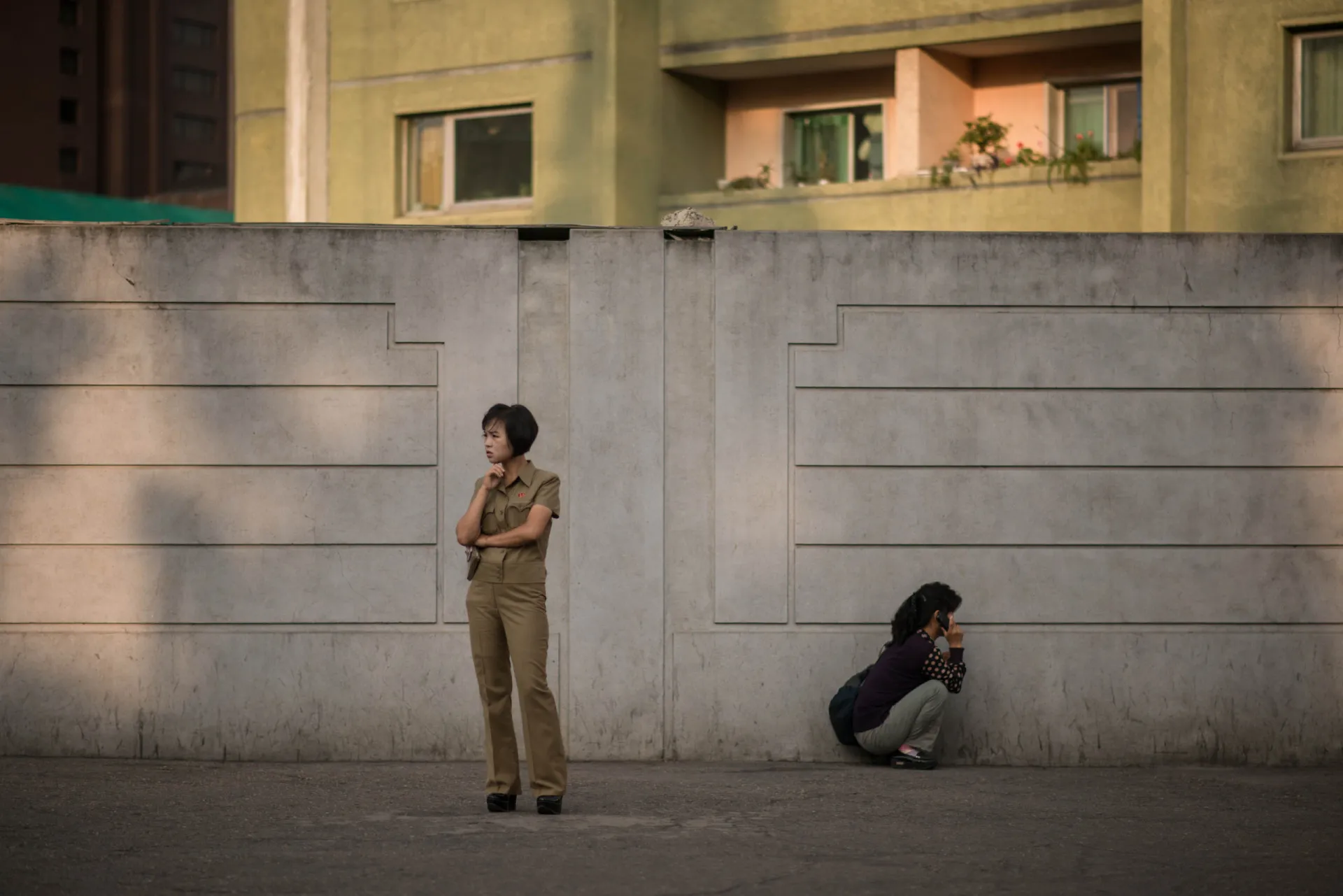Duas pessoas descansando em uma rua em Pyongyang, uma delas de cócoras.