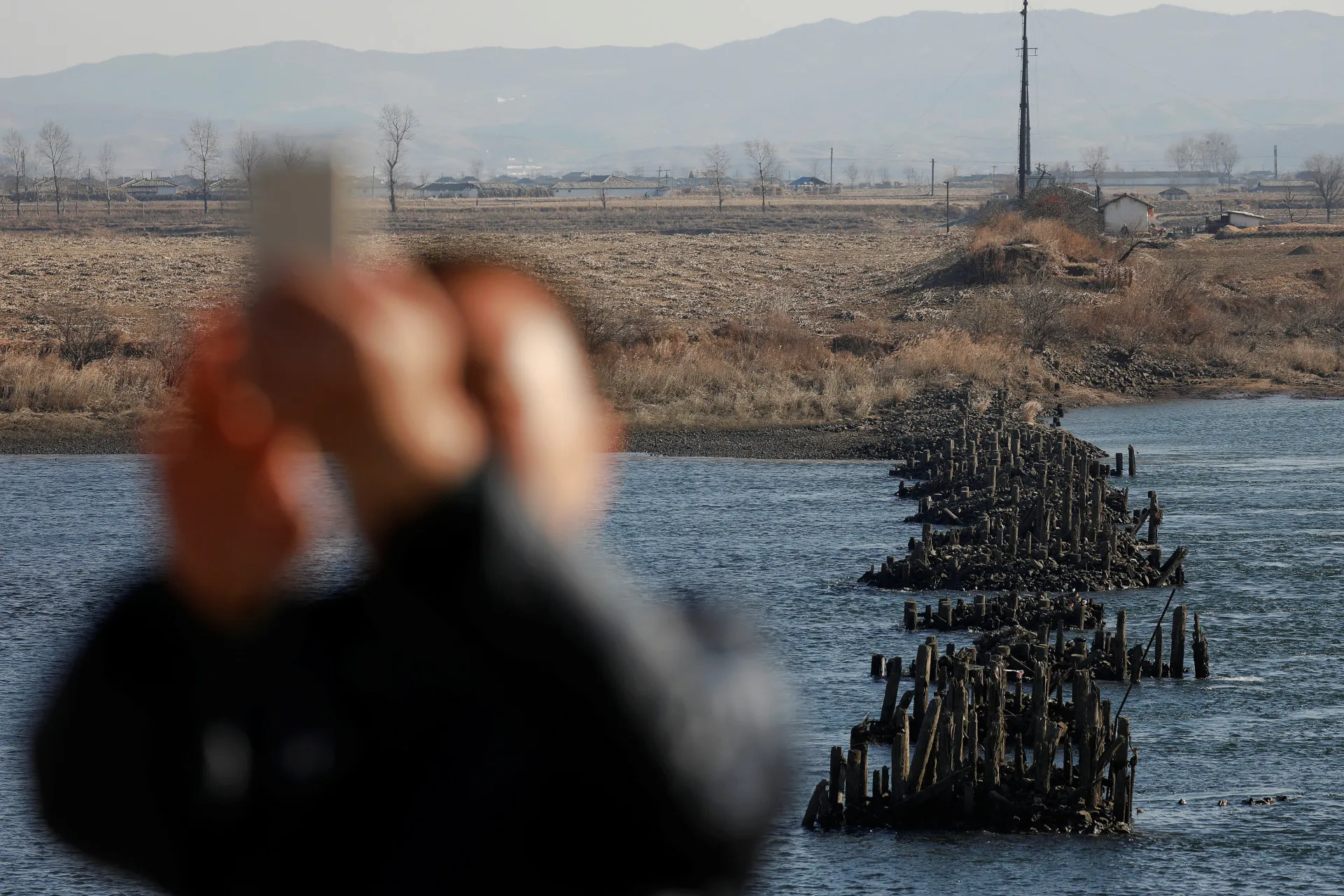 Homem tirando uma selfie com a Coreia do Norte e uma ponte em ruínas ao fundo.