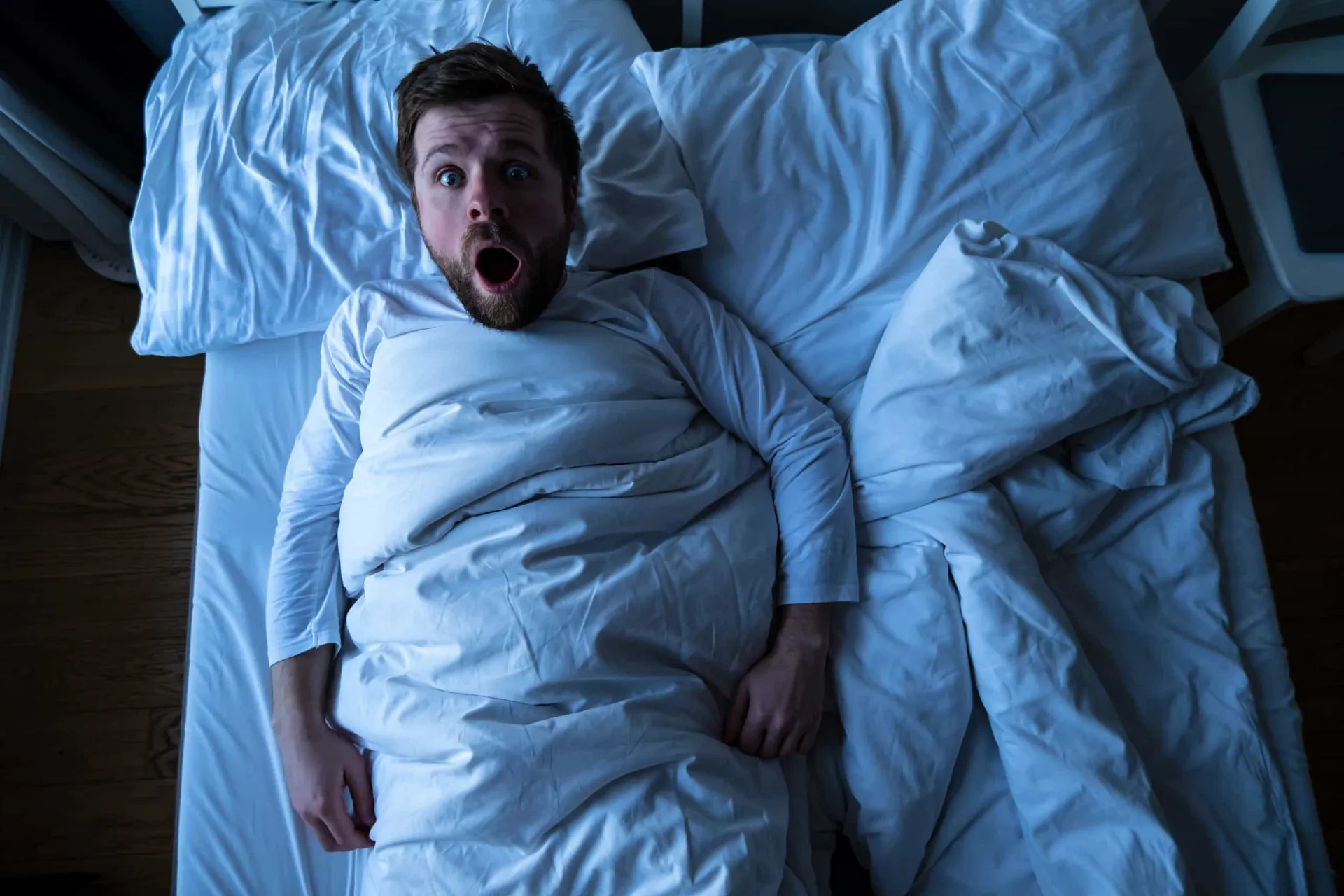 Homem na cama com expressão de terror, como se estivesse tendo um pesadelo.