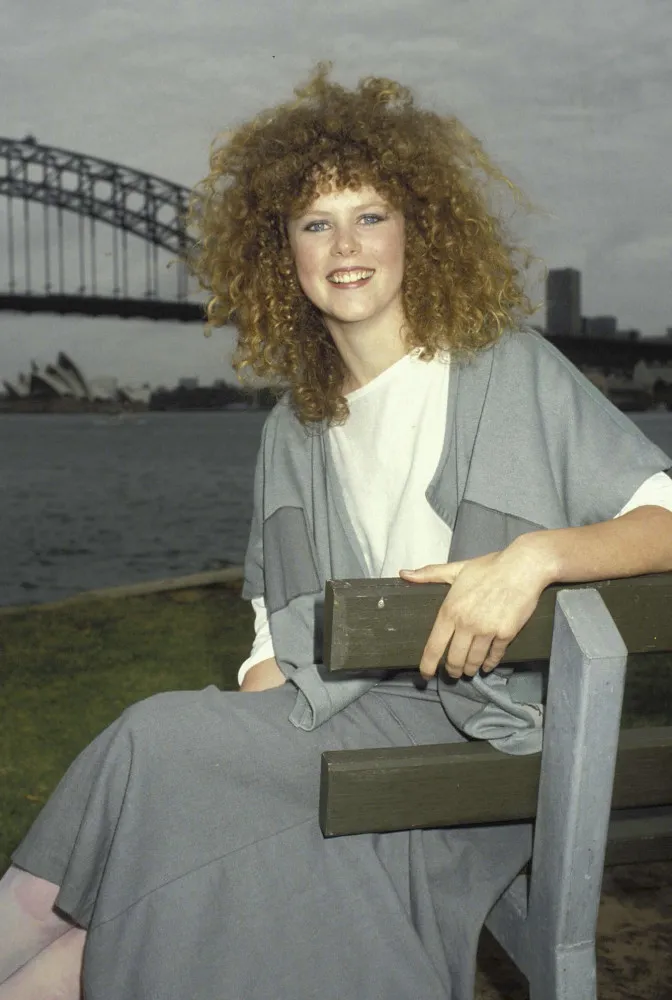 Nicole Kidman em 1983 com cabelo ruivo cacheado e volumoso, vestindo roupas características da década de 80.