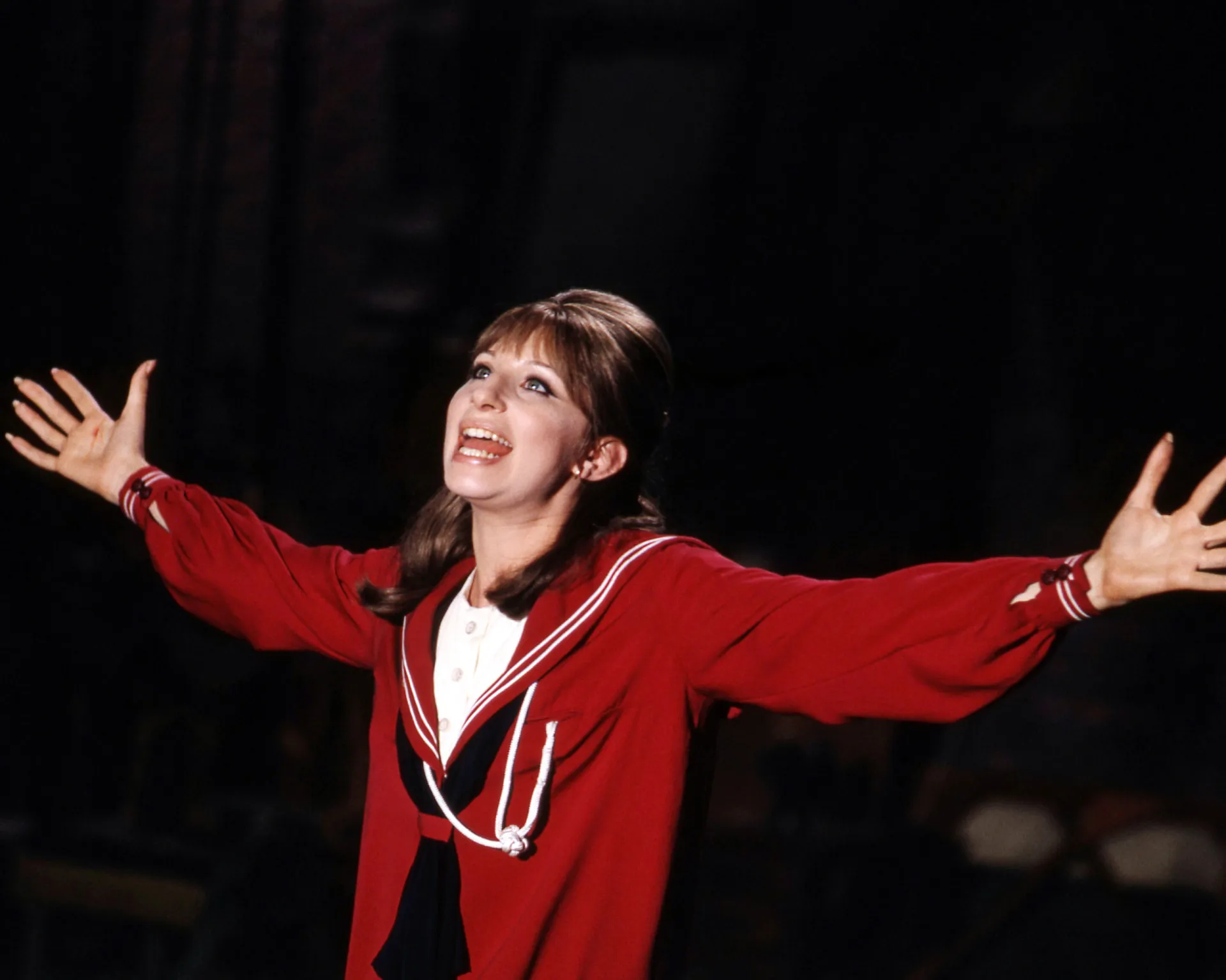 Barbra Streisand cantando intensamente em um palco com microfone na mão.