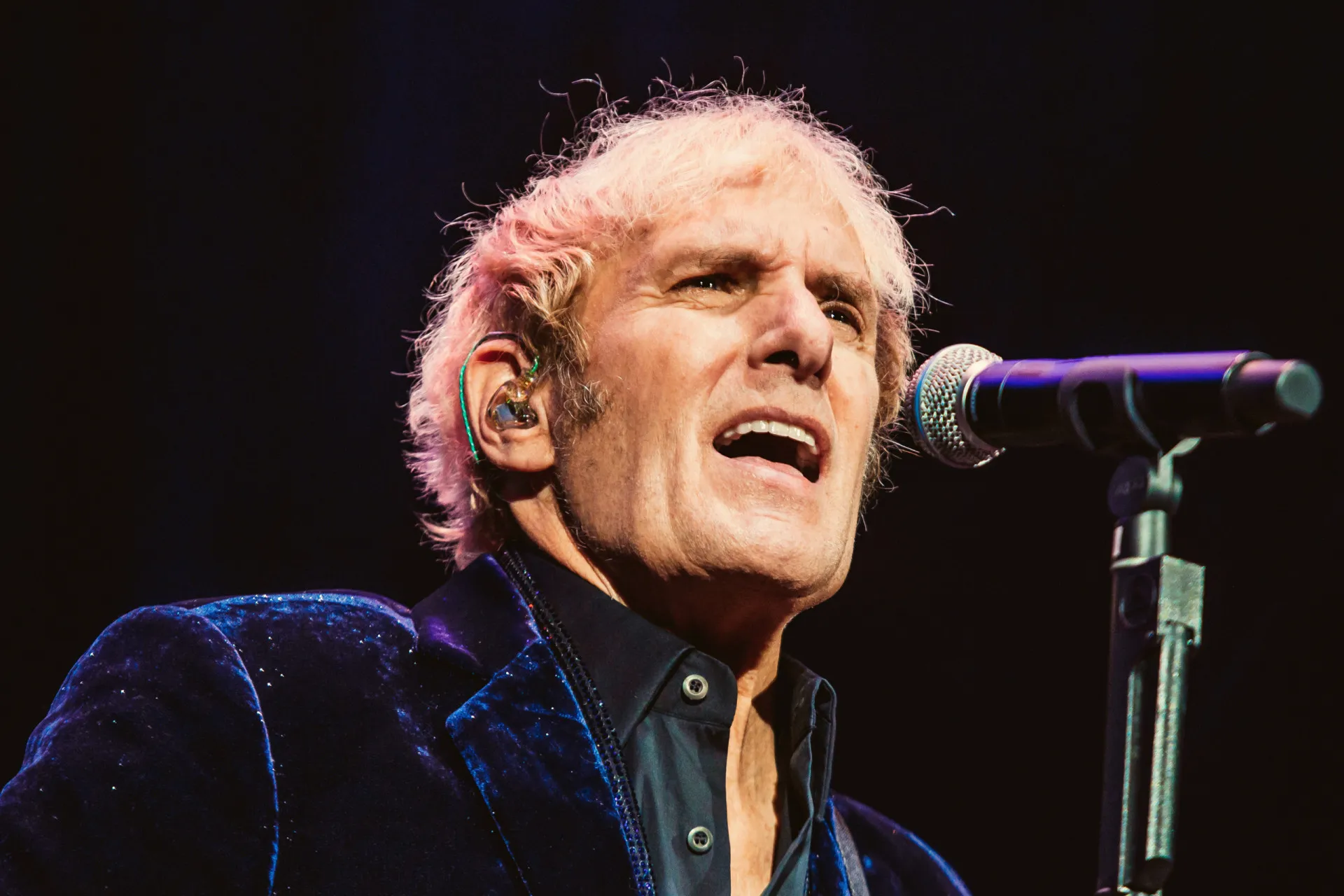 Michael Bolton com cabelos longos, cantando apaixonadamente em um show.