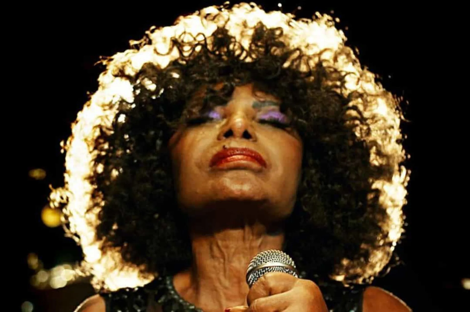A cantora Elza Soares em uma performance vibrante, com maquiagem marcante e expressão poderosa.