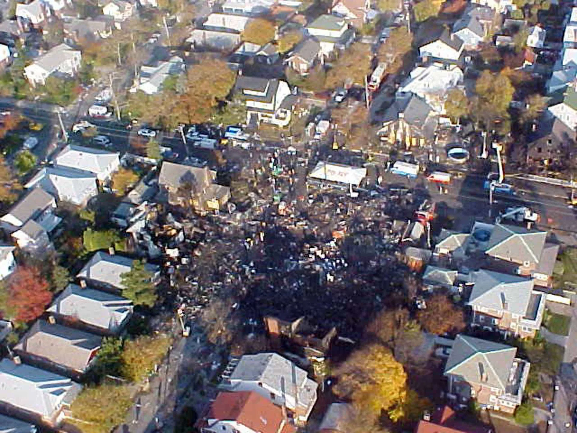 Bombeiros e equipes de emergência em uma rua residencial do Queens, com fumaça subindo dos destroços de um avião.