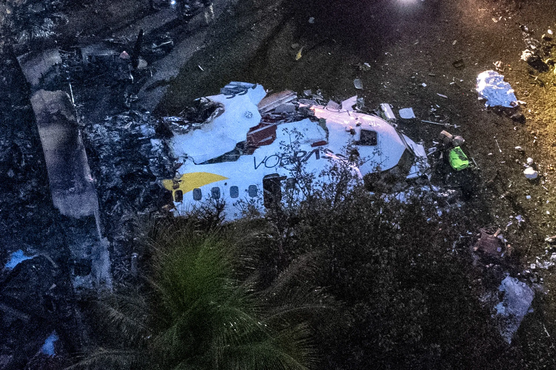 Visão aérea de destroços de avião espalhados por uma área verde, com equipes de investigação no local.
