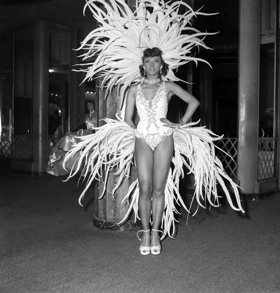 Yvonne Ménard no palco do Folies Bergère, com um sorriso radiante e um figurino luxuoso.