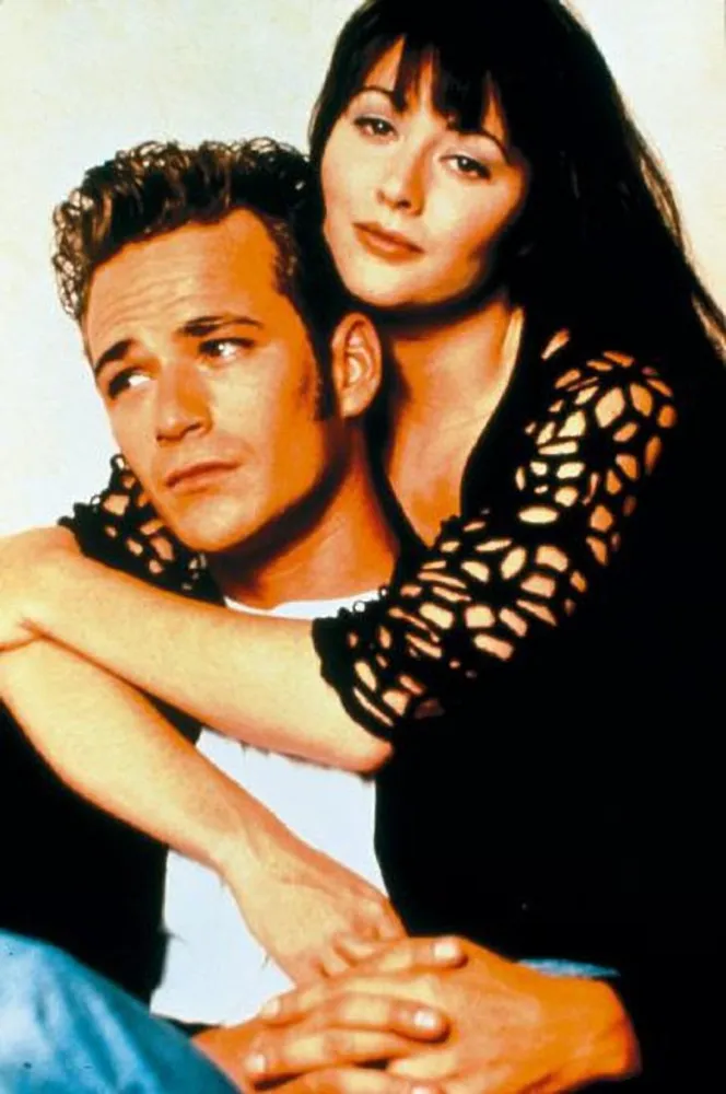 Shannen Doherty como a icônica Brenda Walsh, com seu visual marcante dos anos 90.