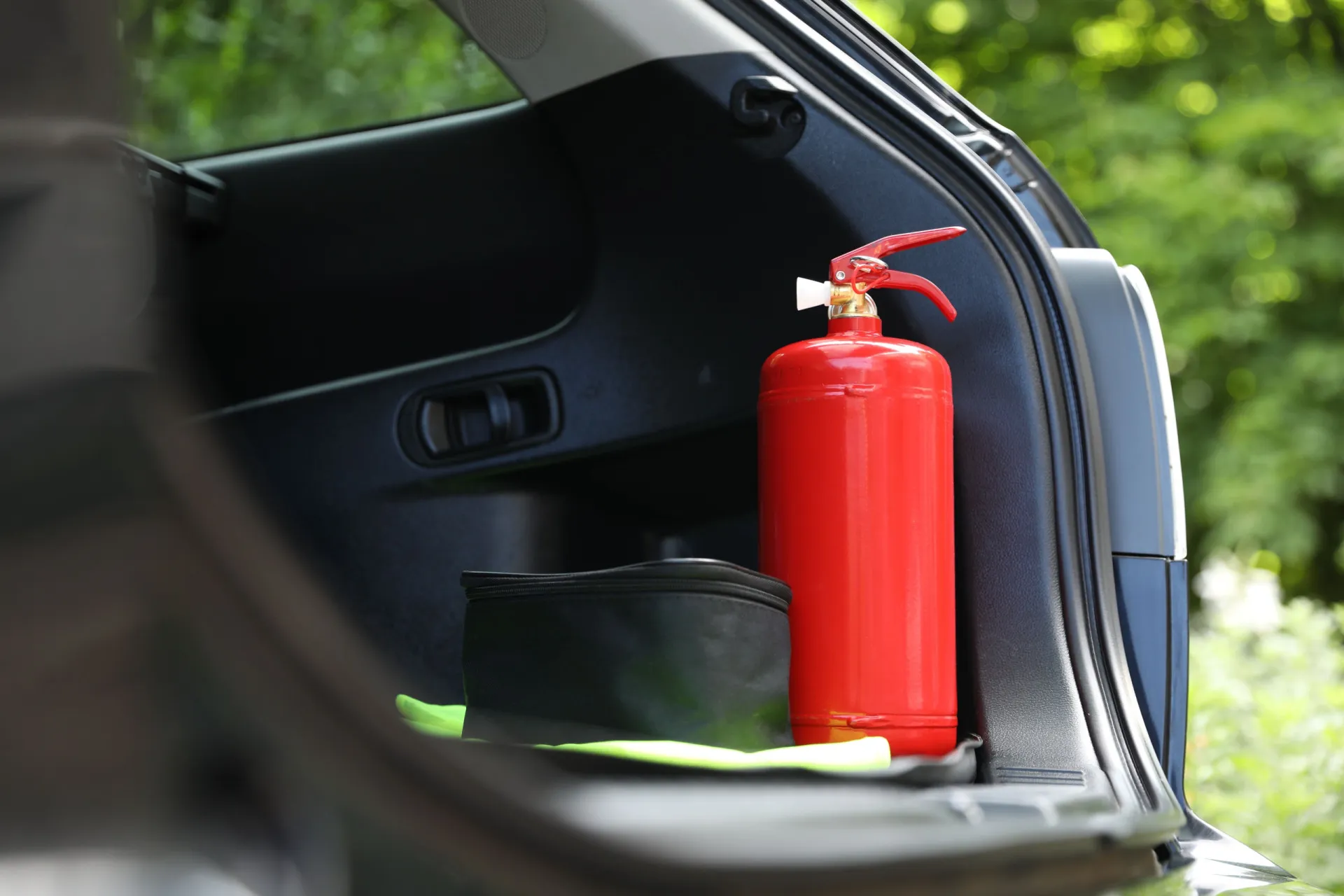 Mão segurando um extintor de incêndio padrão com um carro elétrico em chamas ao fundo, fora de foco.