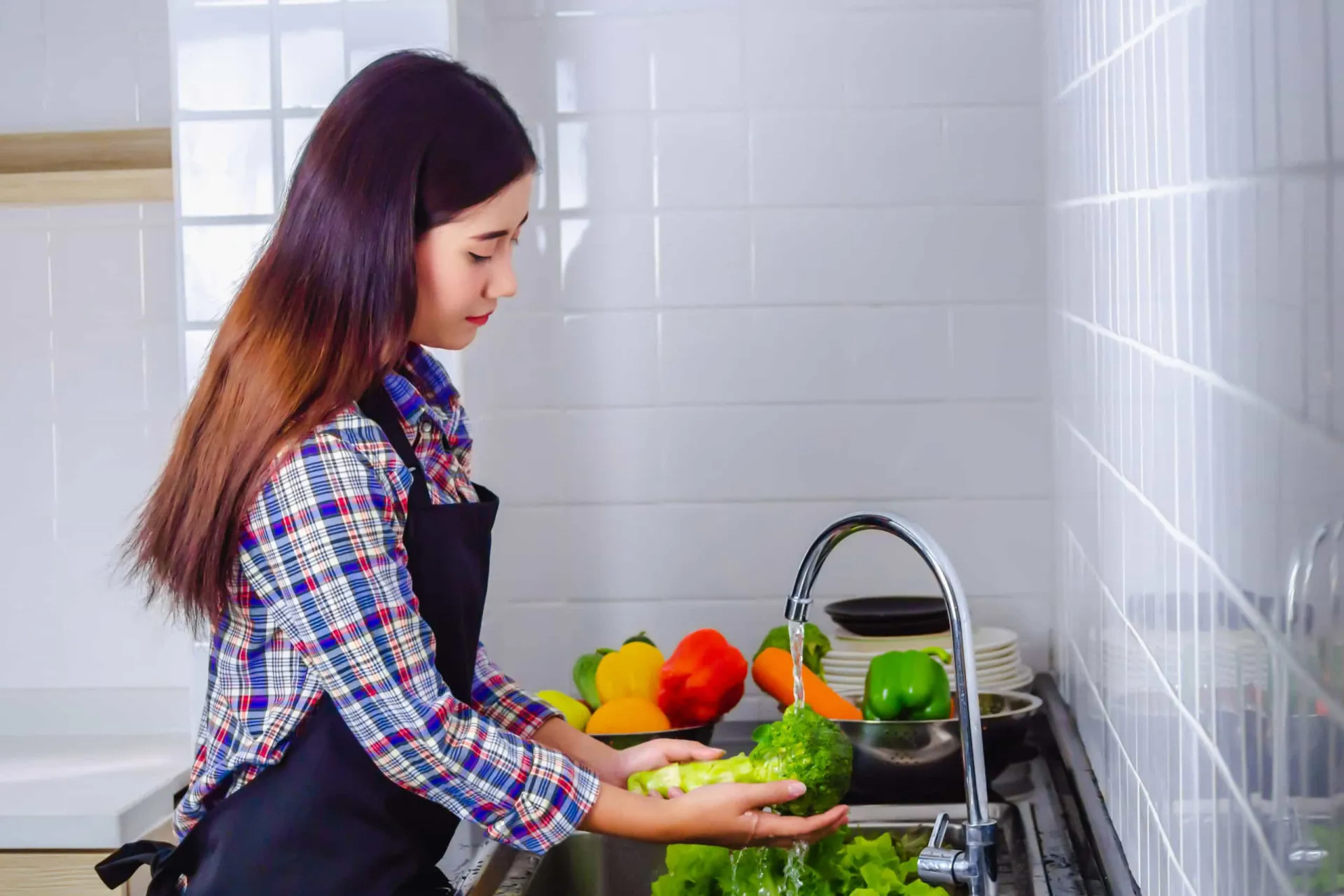 Mãos lavando frutas e vegetais frescos em uma pia com água corrente.