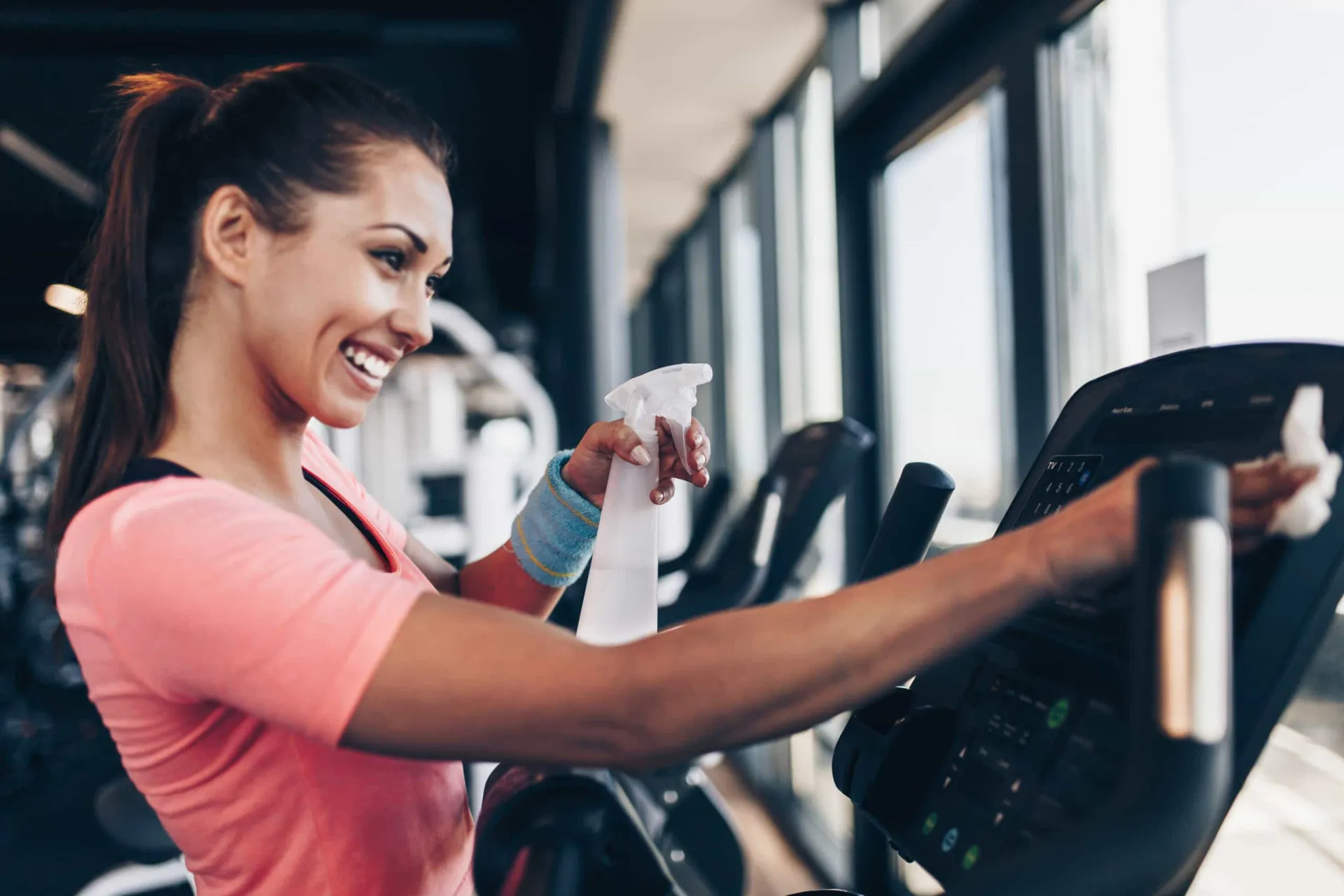 Mulher limpando o assento de um aparelho de musculação com um pano e spray.