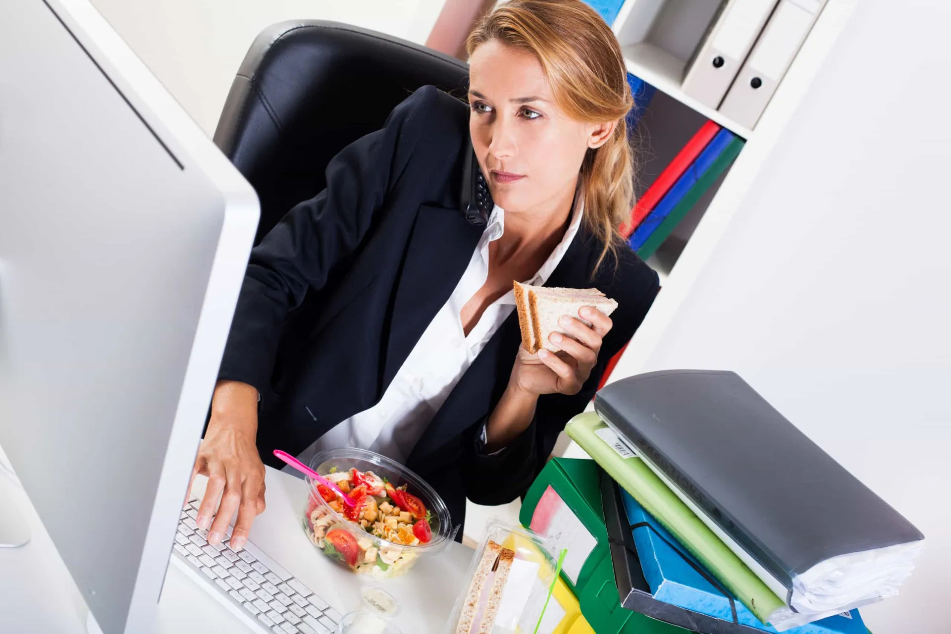 Pessoa comendo um sanduíche enquanto trabalha no notebook em uma mesa de escritório.