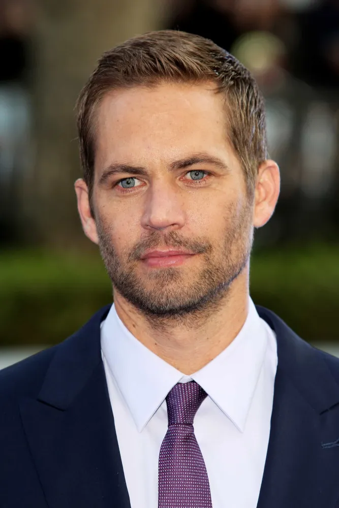 Paul Walker ao lado de um carro esportivo azul, com um olhar sério.
