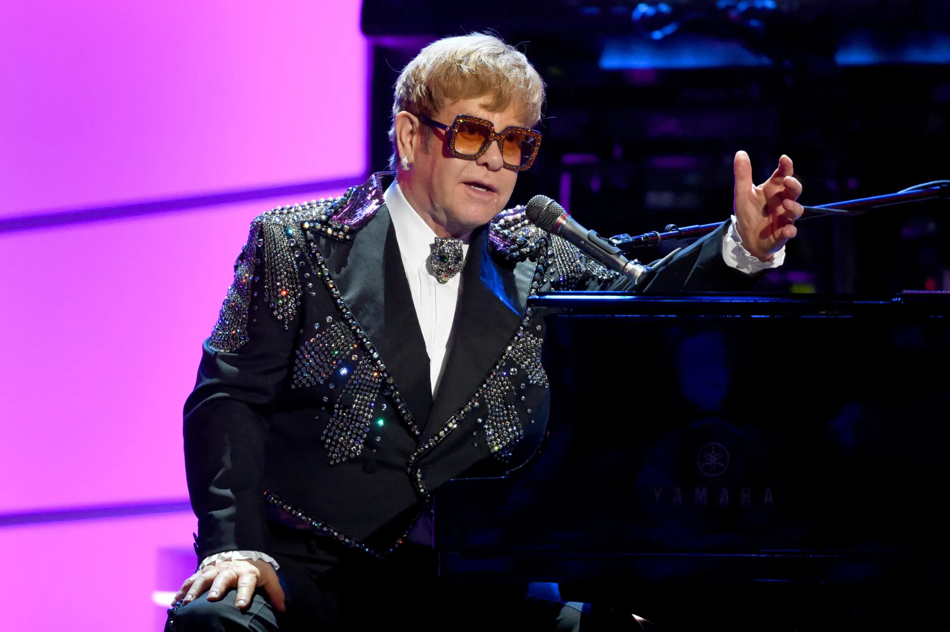 Elton John, com seus óculos icônicos, cantando em um show.