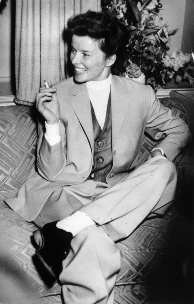 A atriz Katharine Hepburn em uma pose confiante, usando calças e terno, um visual raro para as mulheres de sua época.