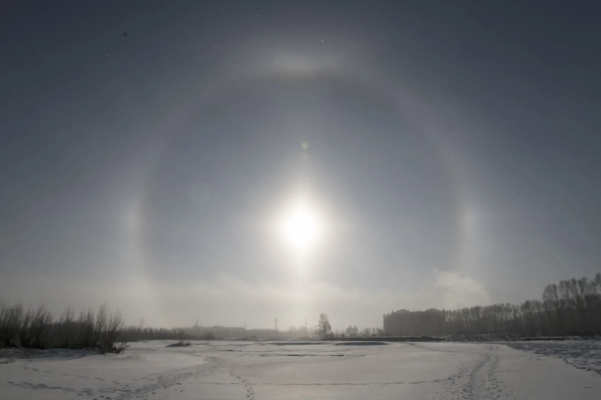 Um grande anel de luz semelhante a um arco-íris circundando o sol no céu, com nuvens finas presentes.