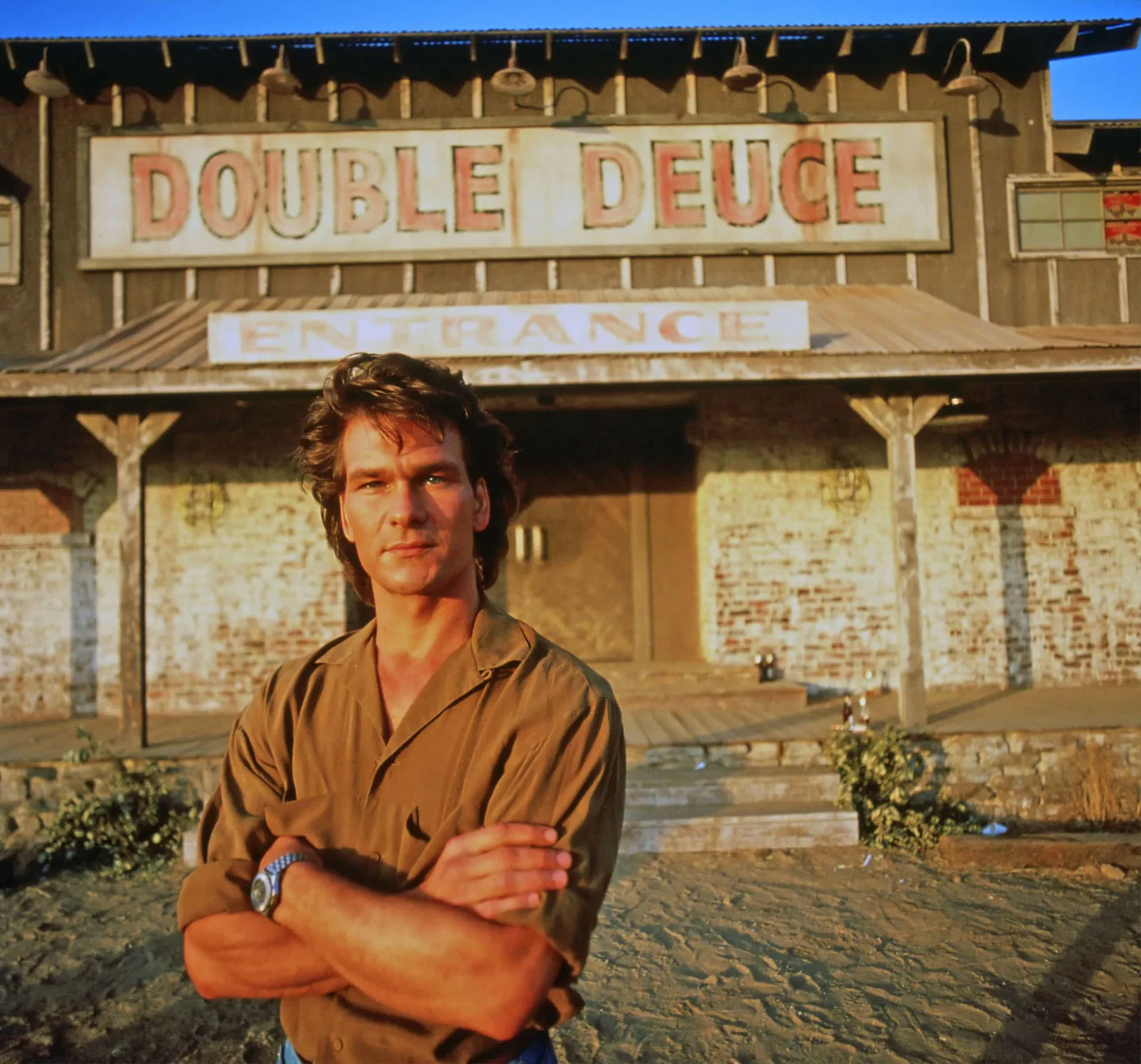 Patrick Swayze como Dalton em 'Matador de Aluguel', com uma pose de durão em um bar.