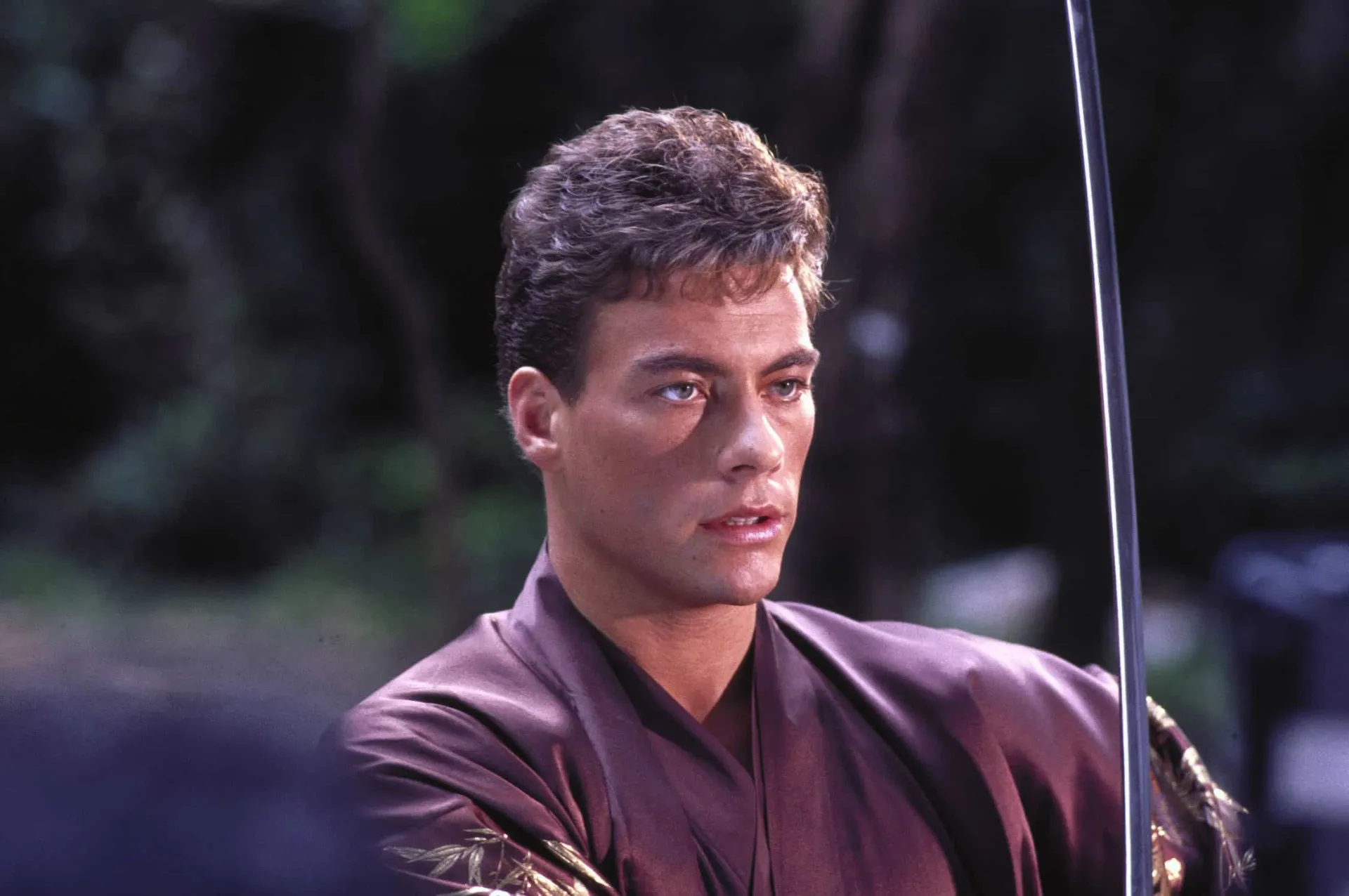 Jean-Claude Van Damme jovem, sem camisa, exibindo seus músculos e fazendo uma pose de luta.