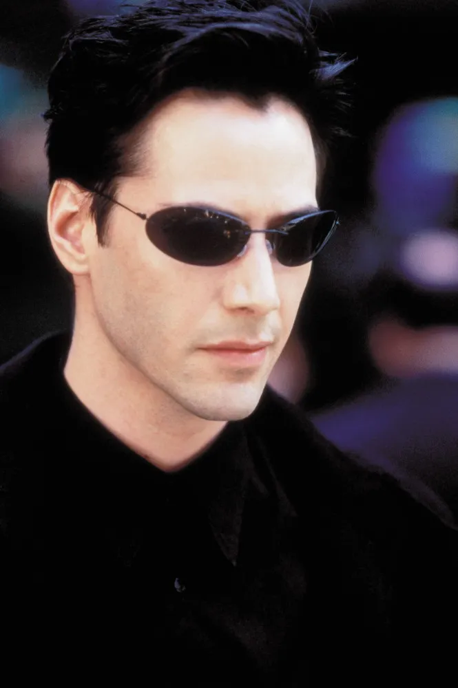 Keanu Reeves jovem como Neo em 'Matrix', com o sobretudo preto e os óculos escuros icônicos.