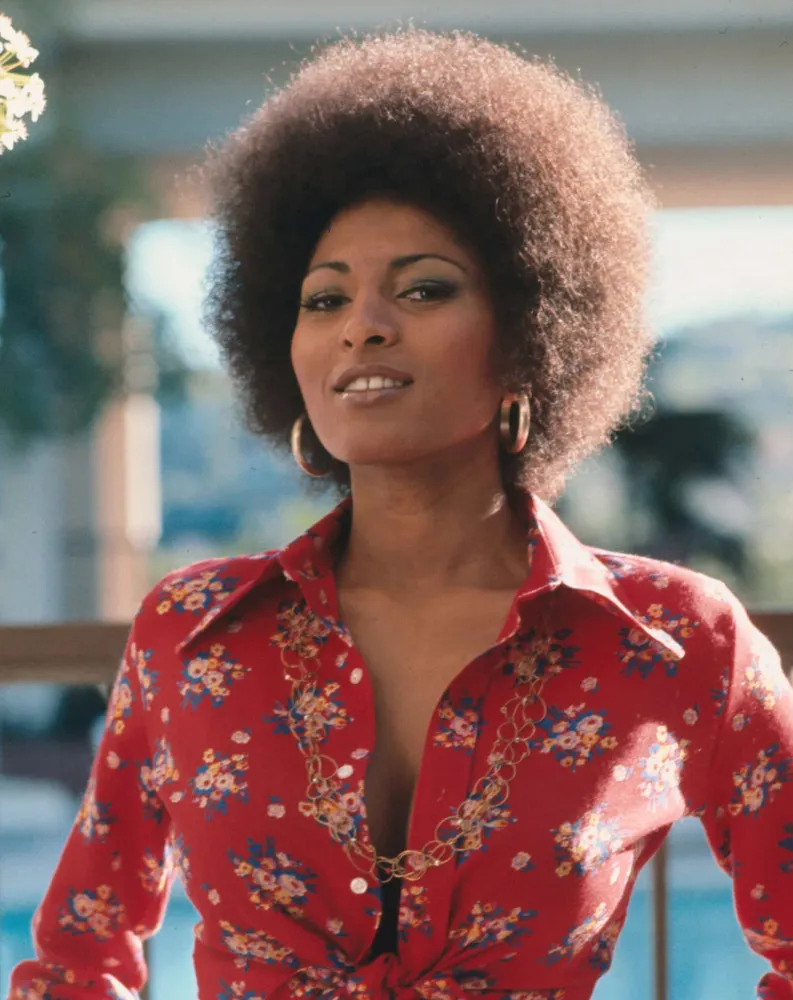 Pam Grier jovem, no auge do movimento Blaxploitation, com seu cabelo afro e olhar poderoso.