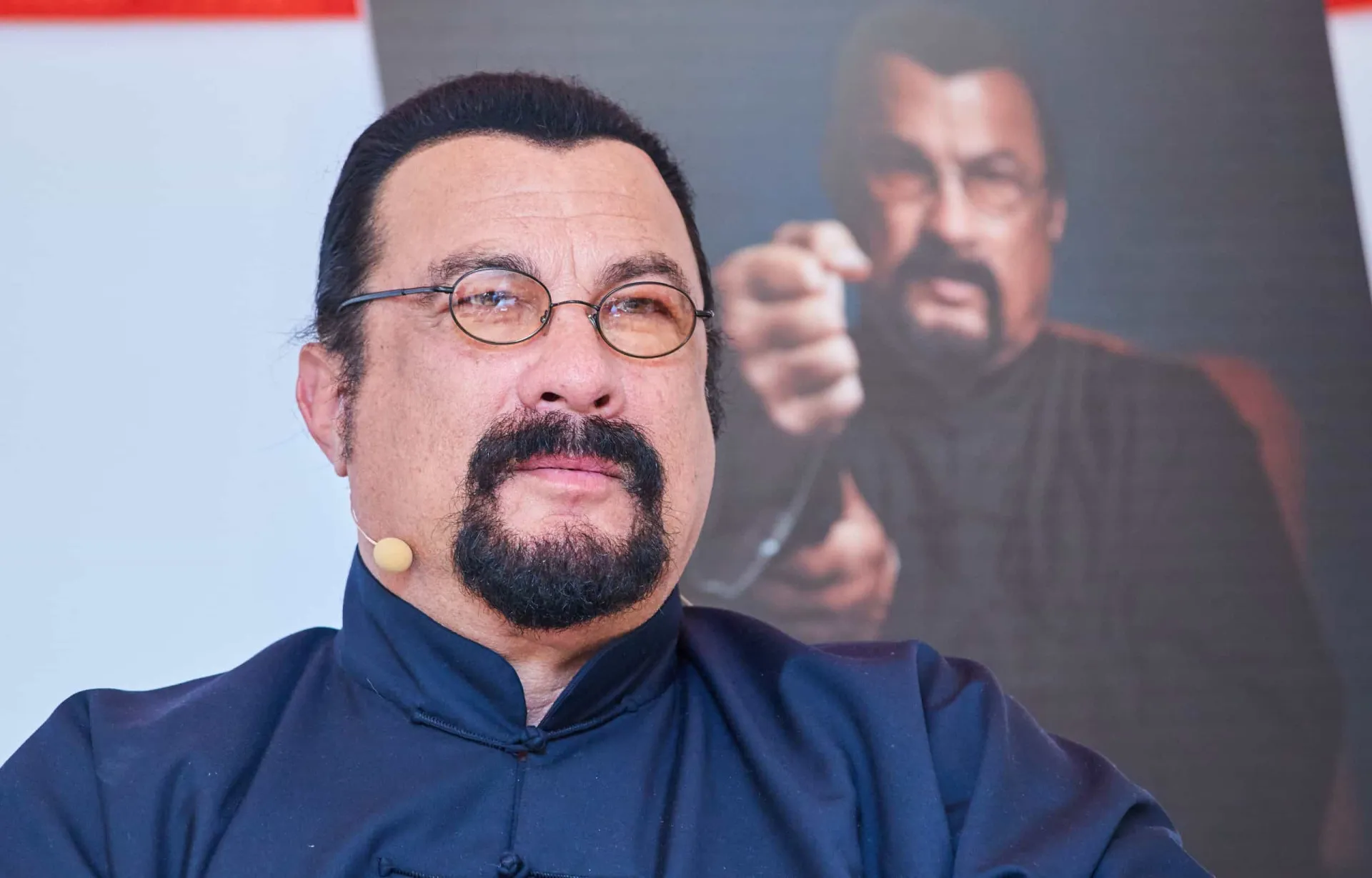 Steven Seagal mais velho, com o rosto mais cheio, cavanhaque e óculos de grau, em um evento público.