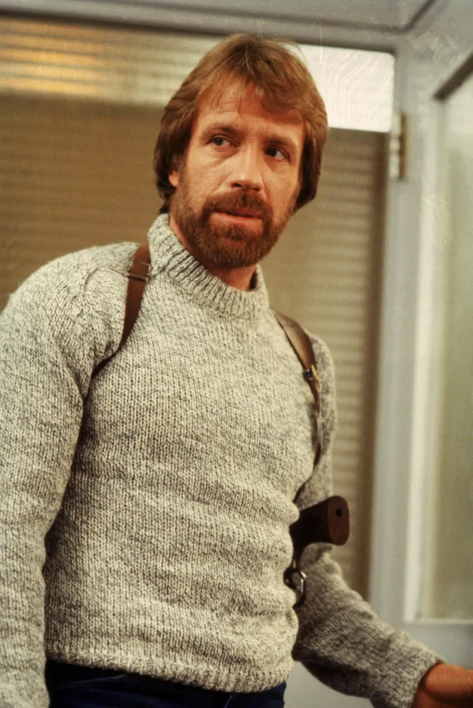 Chuck Norris jovem, nos anos 80, com seu característico bigode e cabelo castanho.