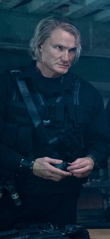 Dolph Lundgren mais velho, com um visual sofisticado de terno, mas ainda com a postura de um astro de ação.