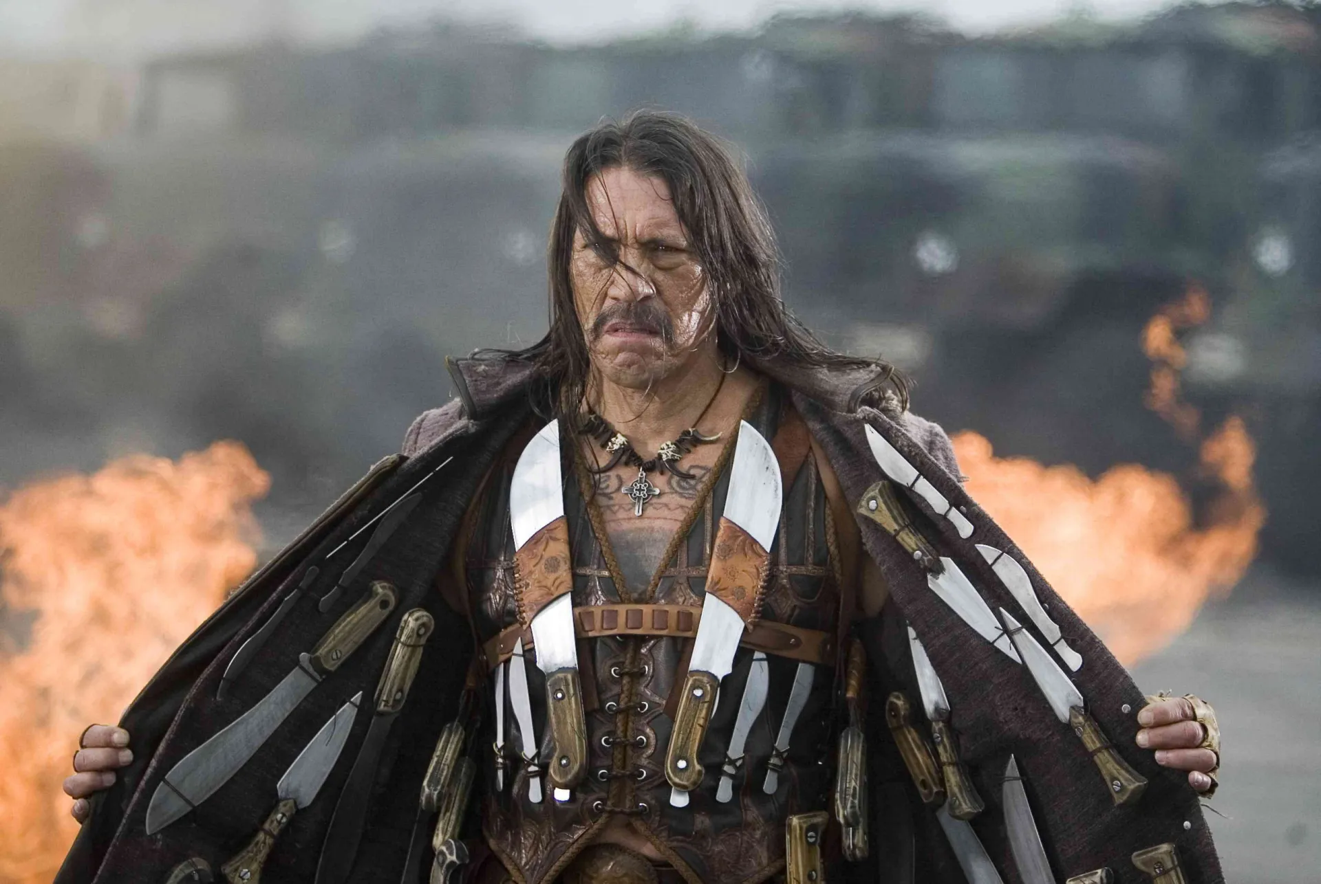 Danny Trejo jovem, com seu longo cabelo preto e bigode característico, exibindo as tatuagens no peito.