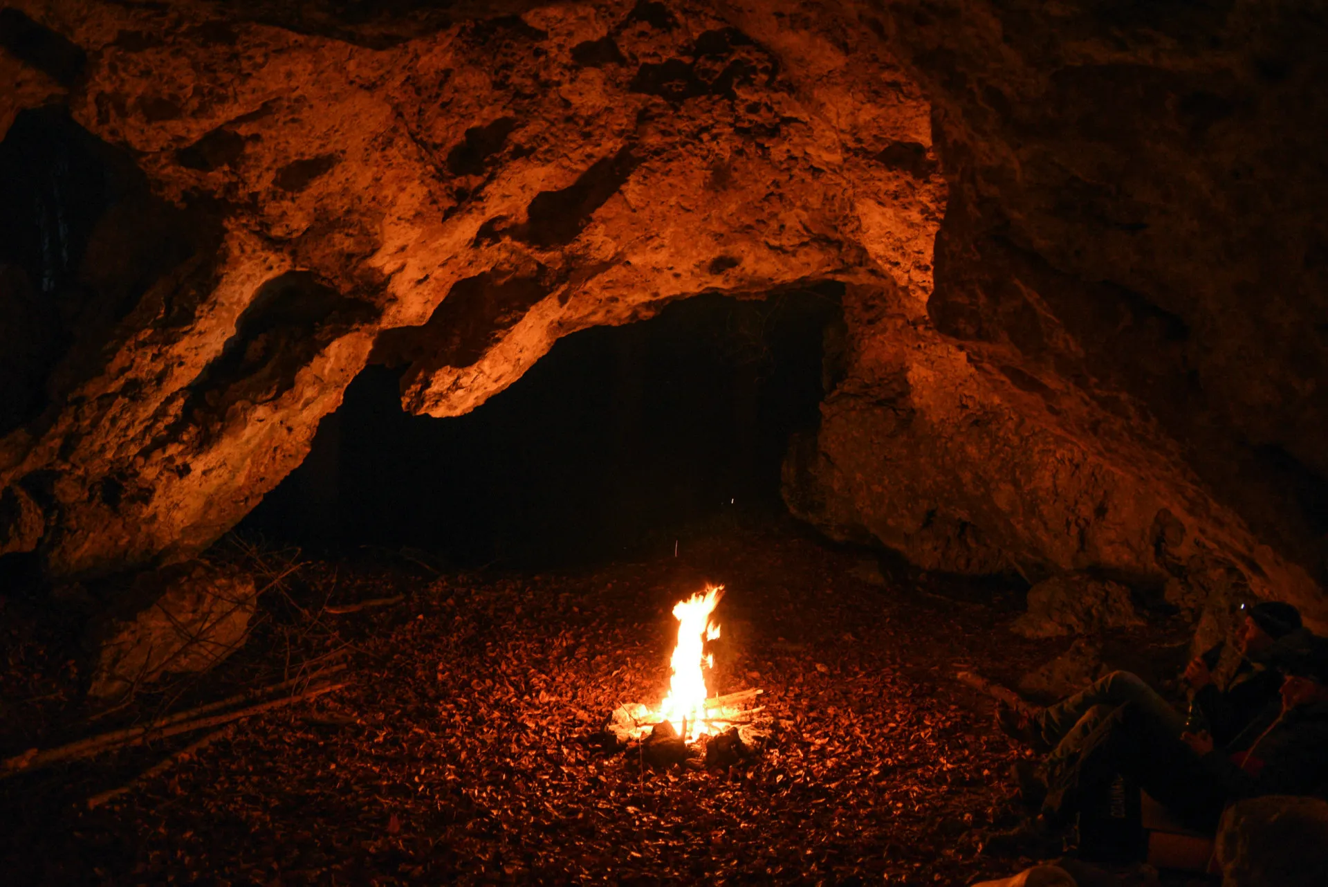 Pessoa fazendo uma fogueira na entrada de uma caverna escura.