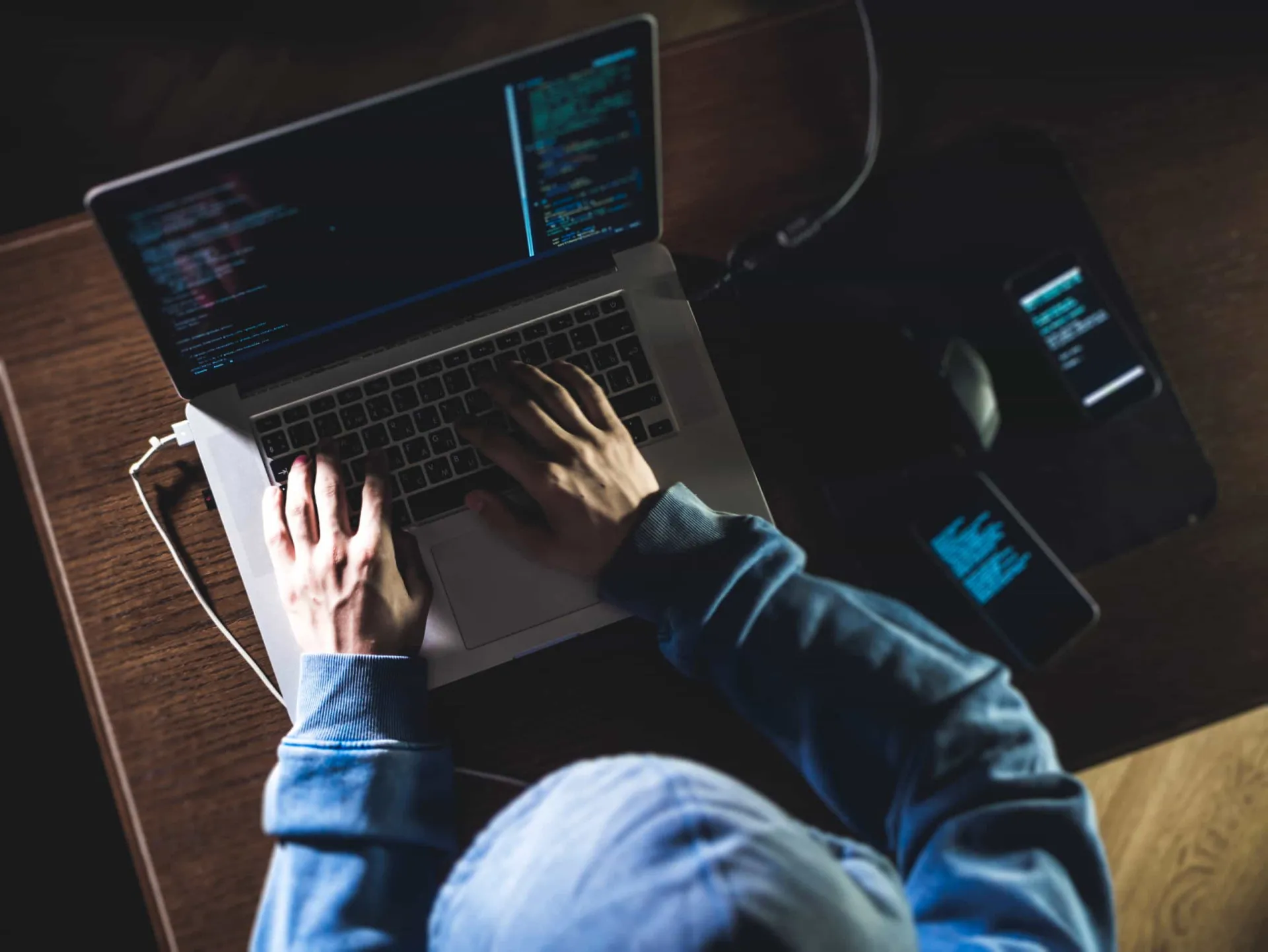 Pessoa com capuz digitando muito rápido em um laptop em um ambiente escuro, representando um hacker de filme.