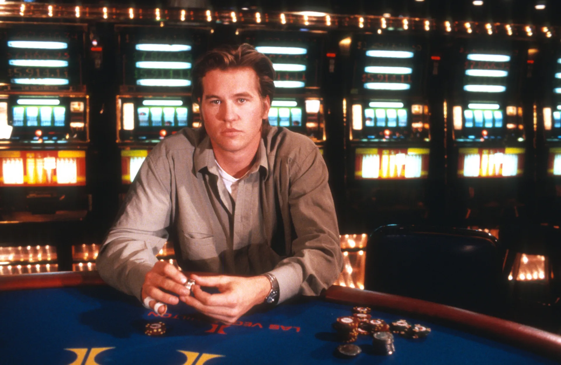 O ator Val Kilmer com seu visual icônico dos anos 90, olhando intensamente para a câmera.