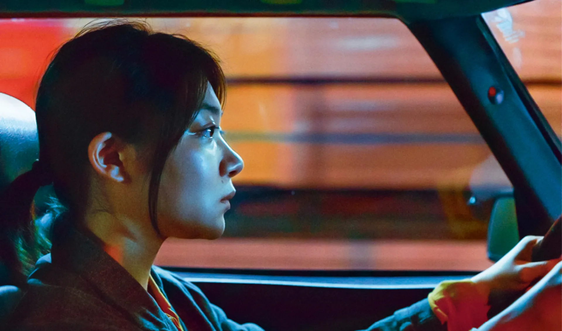 Cena do filme 'Drive My Car', mostrando os personagens dentro de um carro vermelho.