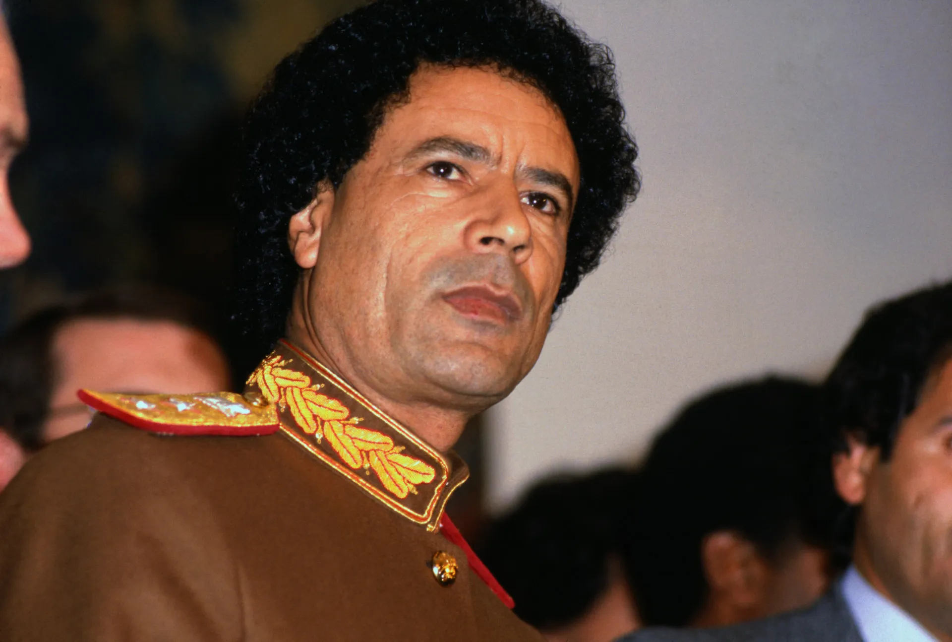 Muammar al-Gaddafi com seus trajes característicos e óculos de sol, parecendo enigmático.