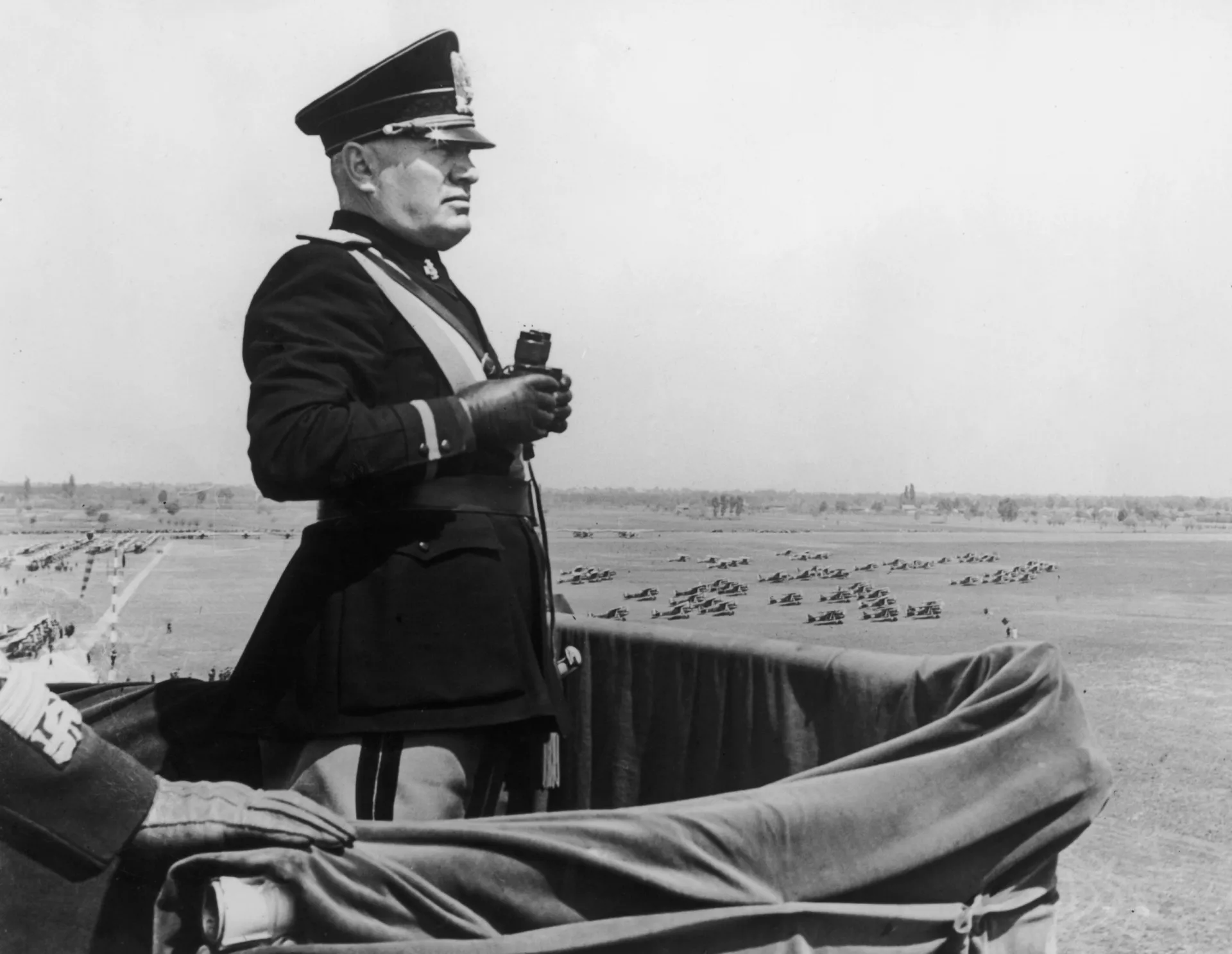Benito Mussolini discursando para uma multidão, com expressão enérgica e gesto imponente.