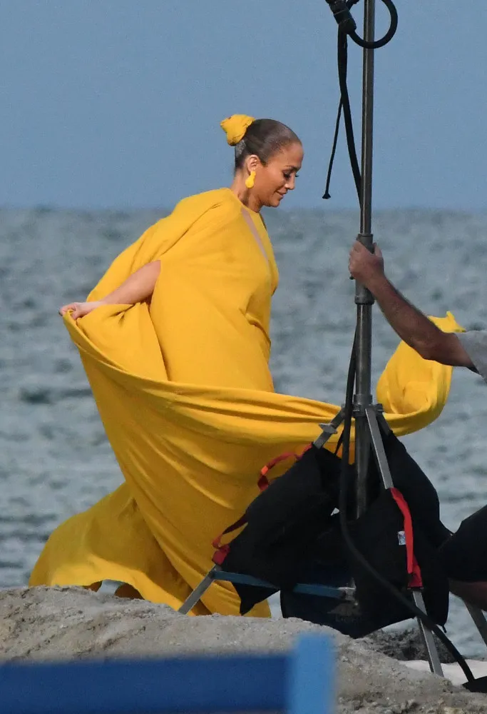 Jennifer Lopez segurando seu longo vestido amarelo que é levantado com força pelo vento durante a gravação de um clipe.